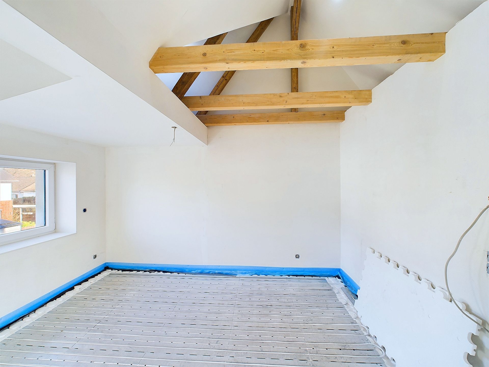 Heller Raum mit sichtbaren Holzbalken und unfertigem Boden, weiß gestrichene Wände, Fenster links.