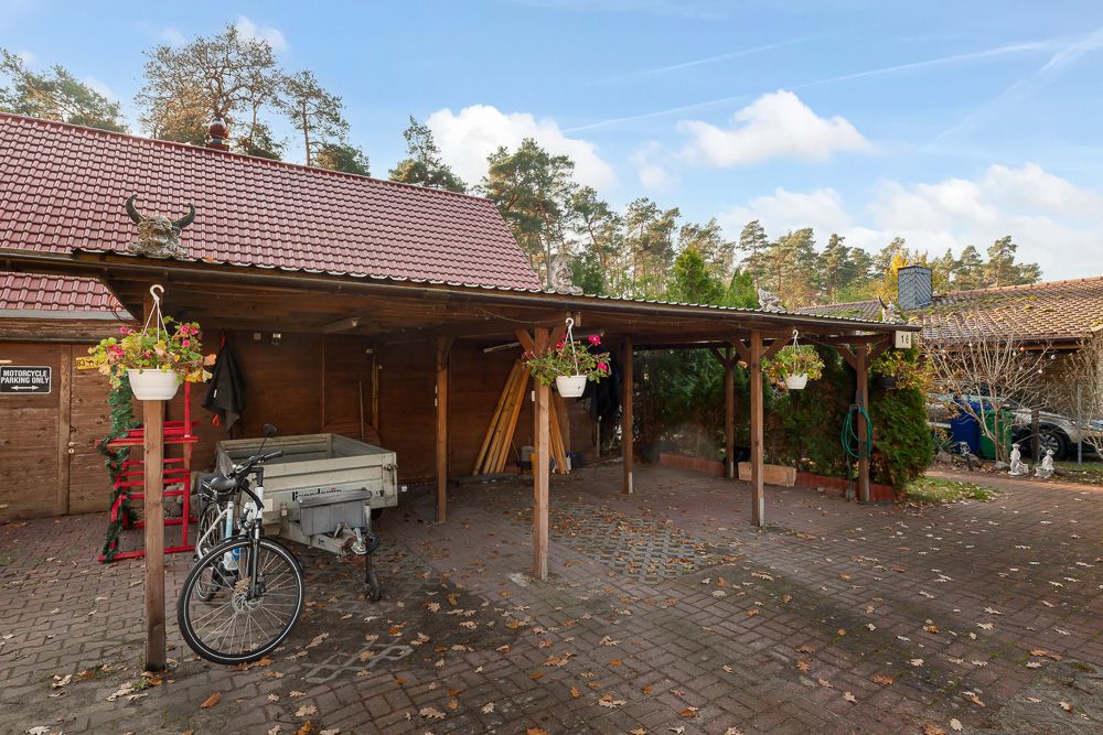 Holz-Carport mit Blumen, Fahrrad, Anhänger und Pflastersteinen vor einem Haus im Grünen.