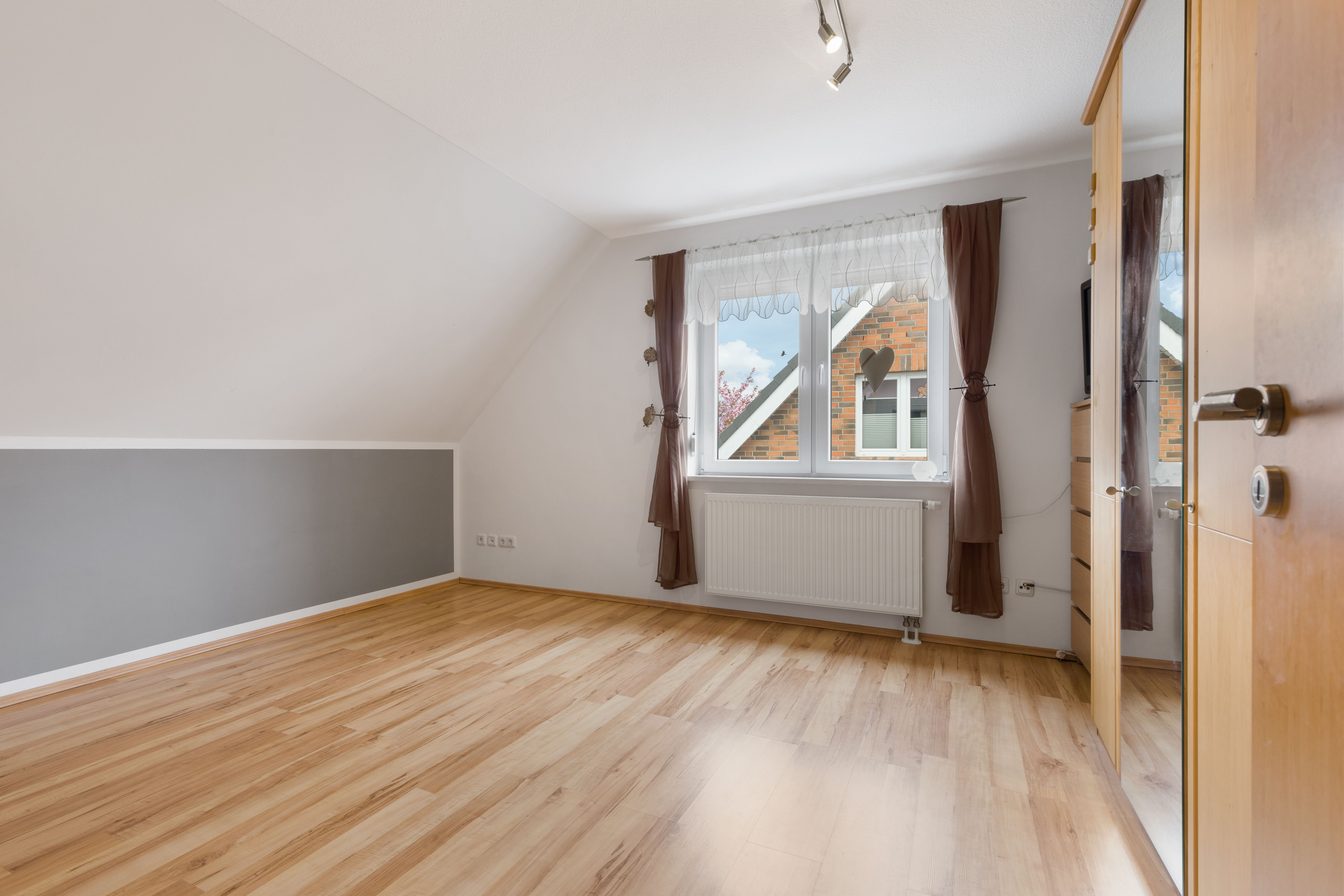 Helles Zimmer mit Parkettboden, großem Fenster, braunen Vorhängen und Einbauschrank.