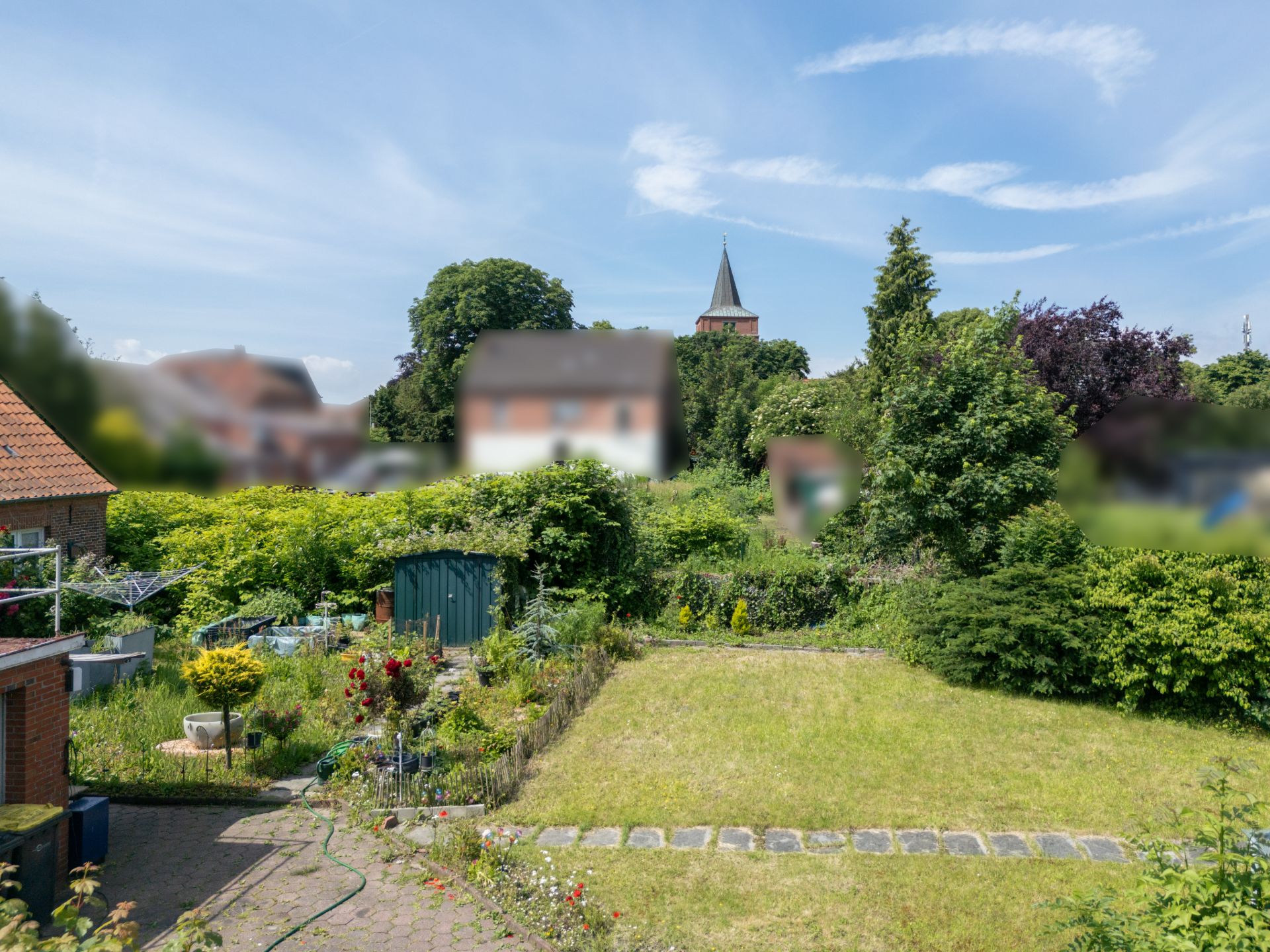 Blick auf einen grünen Garten mit Rasenfläche, Beeten und Gartenhaus, umgeben von Büschen.