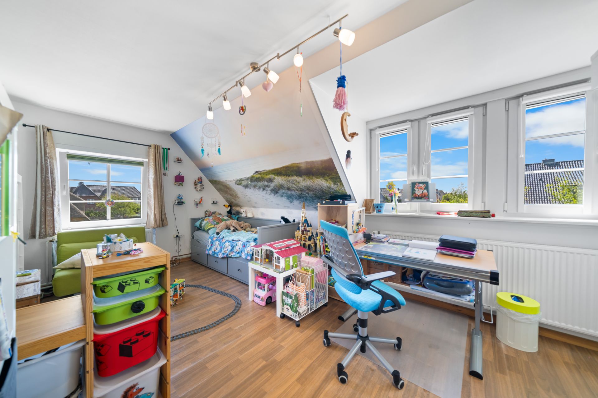 Helles Kinderzimmer mit Schreibtisch, Spielzeugregalen und Wandbild einer Strandlandschaft.