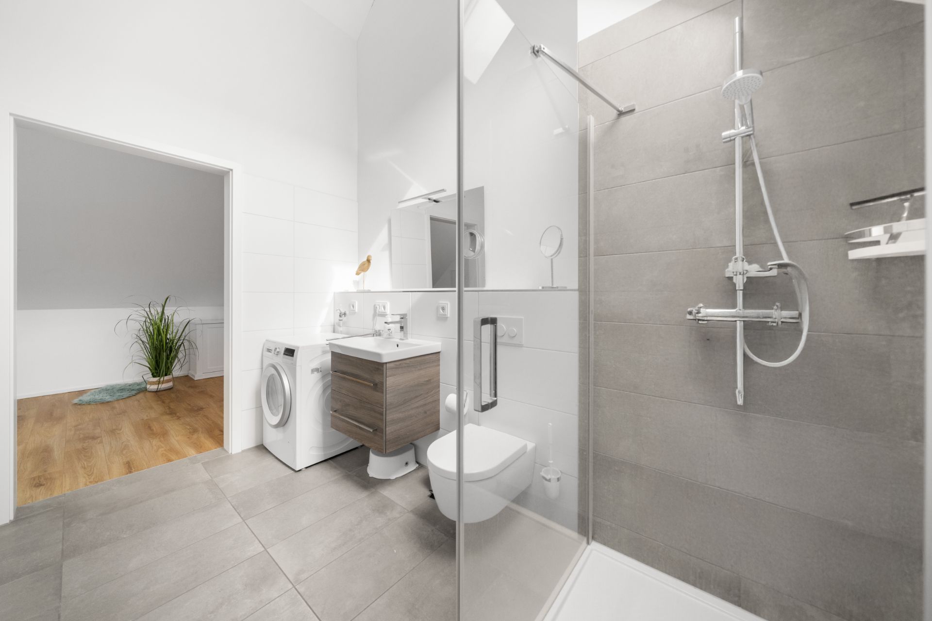 Modernes Badezimmer mit Dusche, Waschbecken, Toilette und Waschmaschine, angrenzend an einen Raum mit Holzboden.