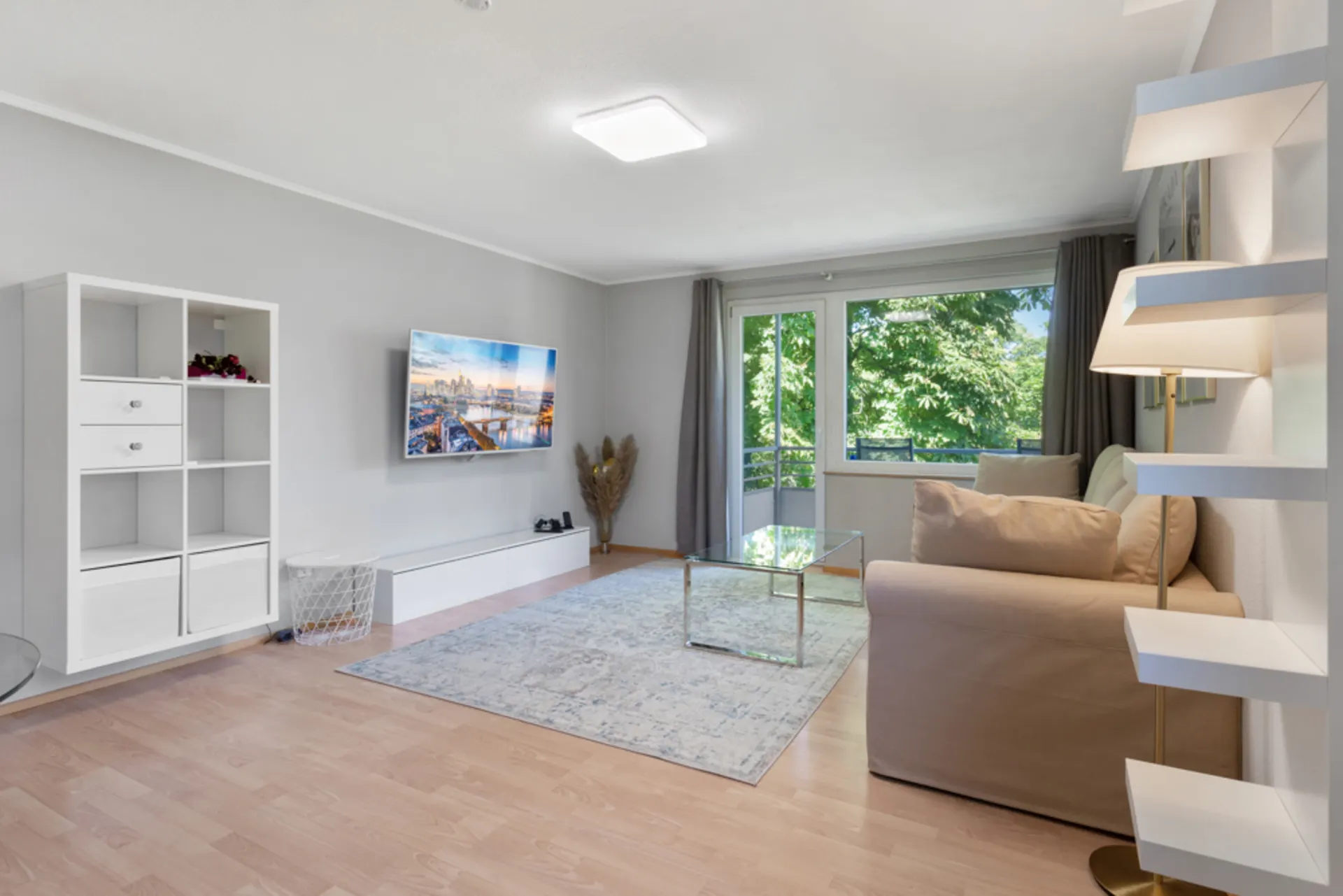 Helles Wohnzimmer mit Sofa, Glastisch, Regal, Fernseher und großem Fenster mit Blick ins Grüne.