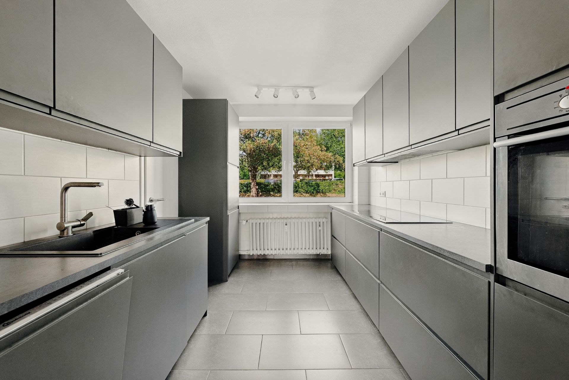 Moderne Küche mit grauen Schränken, schwarzen Arbeitsflächen und Fenstern mit Blick ins Grüne.