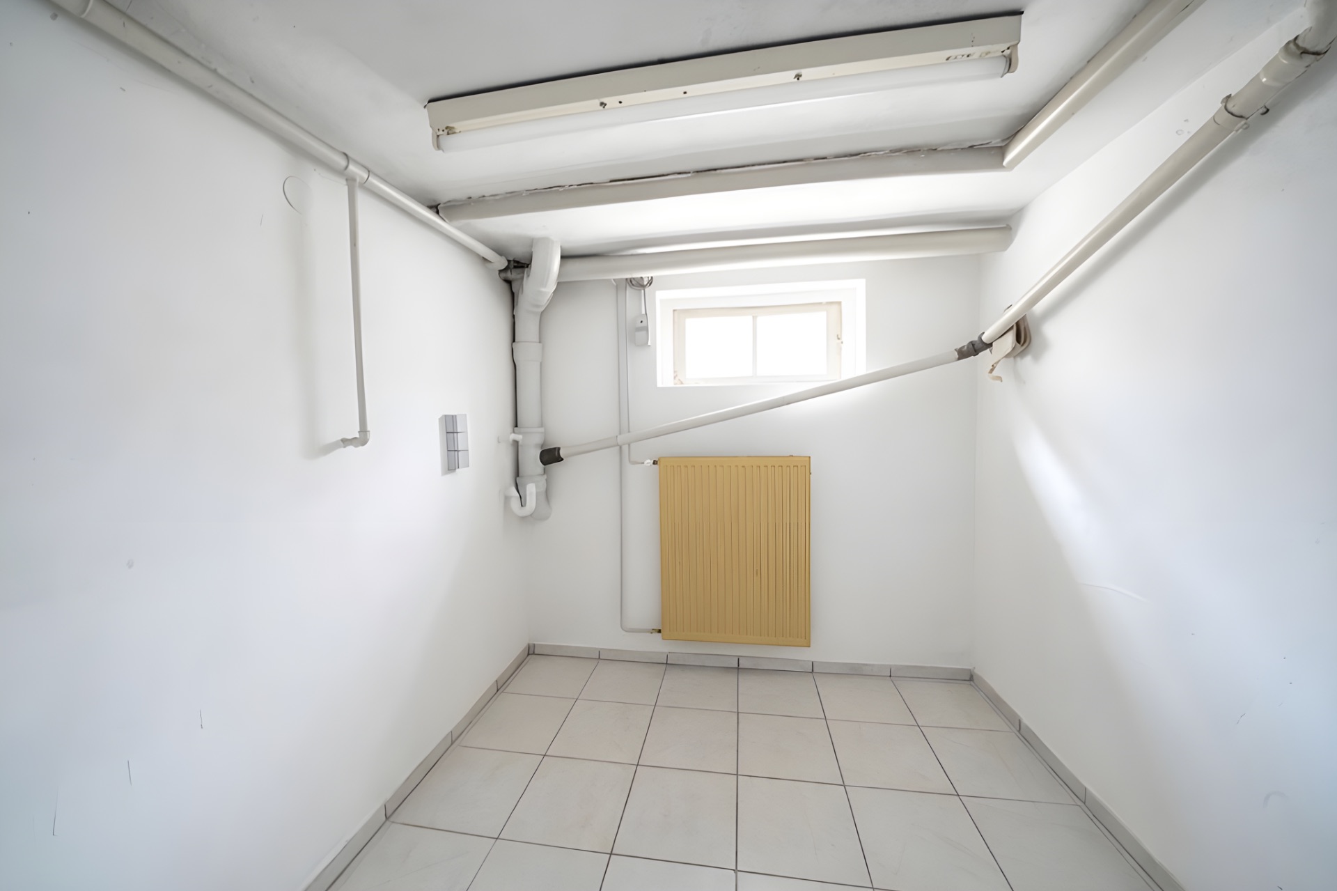 Kleiner weißer Raum mit Fliesenboden, gelbem Heizkörper und sichtbaren Rohren an den Wänden.