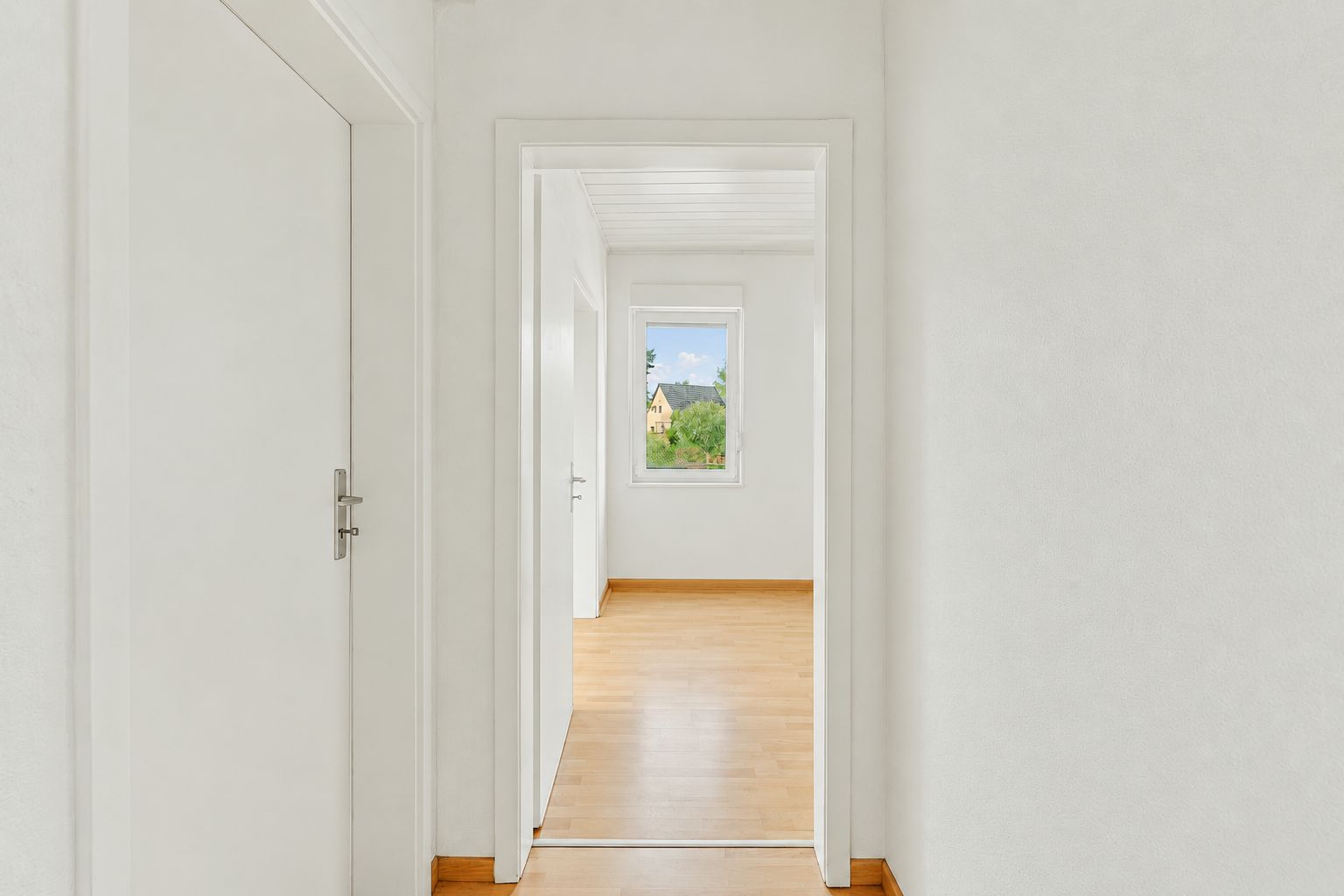 Heller Flur mit Blick in ein leeres Zimmer mit Fenster und Parkettboden.