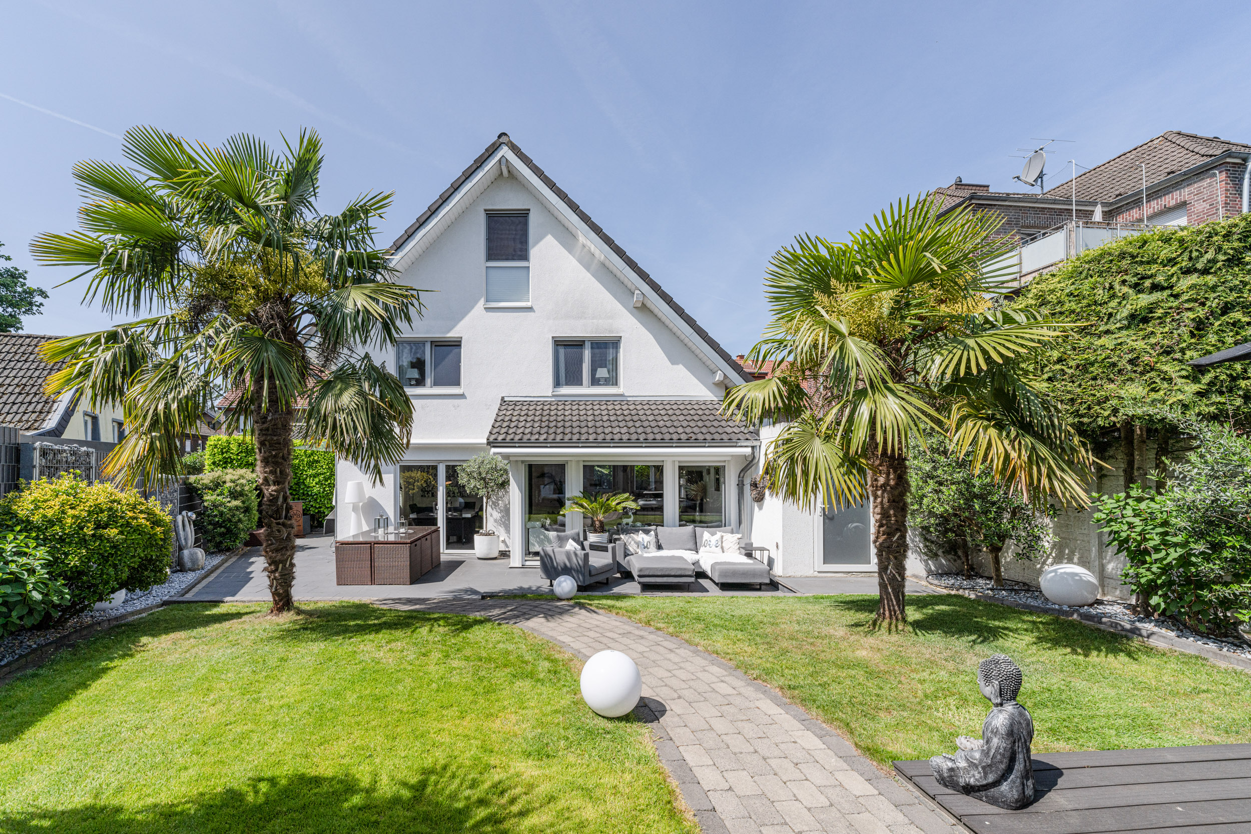 Gepflegter Garten mit Palmen, Rasen, Gehweg und Sitzecke vor einem weißen Einfamilienhaus.