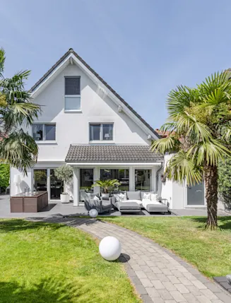 Gepflegter Garten mit Palmen, Rasen, Gehweg und Sitzecke vor einem weißen Einfamilienhaus.