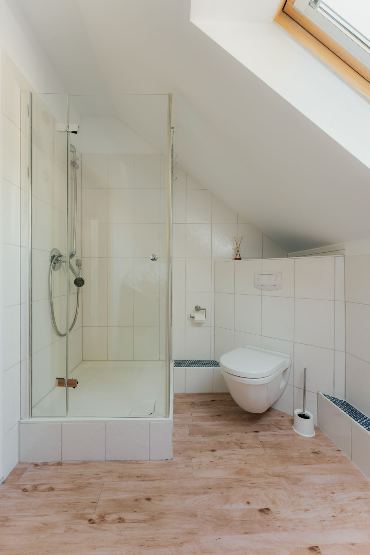 Modernes Badezimmer mit Dusche, Toilette und Dachfenster, hell gefliest und mit Holzfußboden.