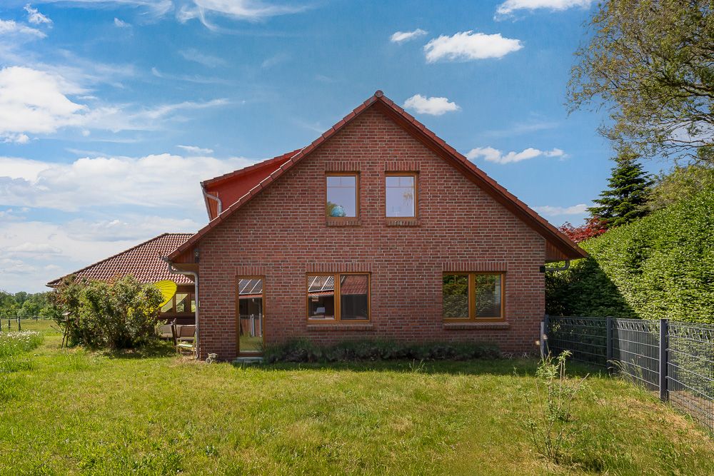 Backsteinwohnhaus mit Satteldach und Garten, umgeben von Hecken und Zaun.