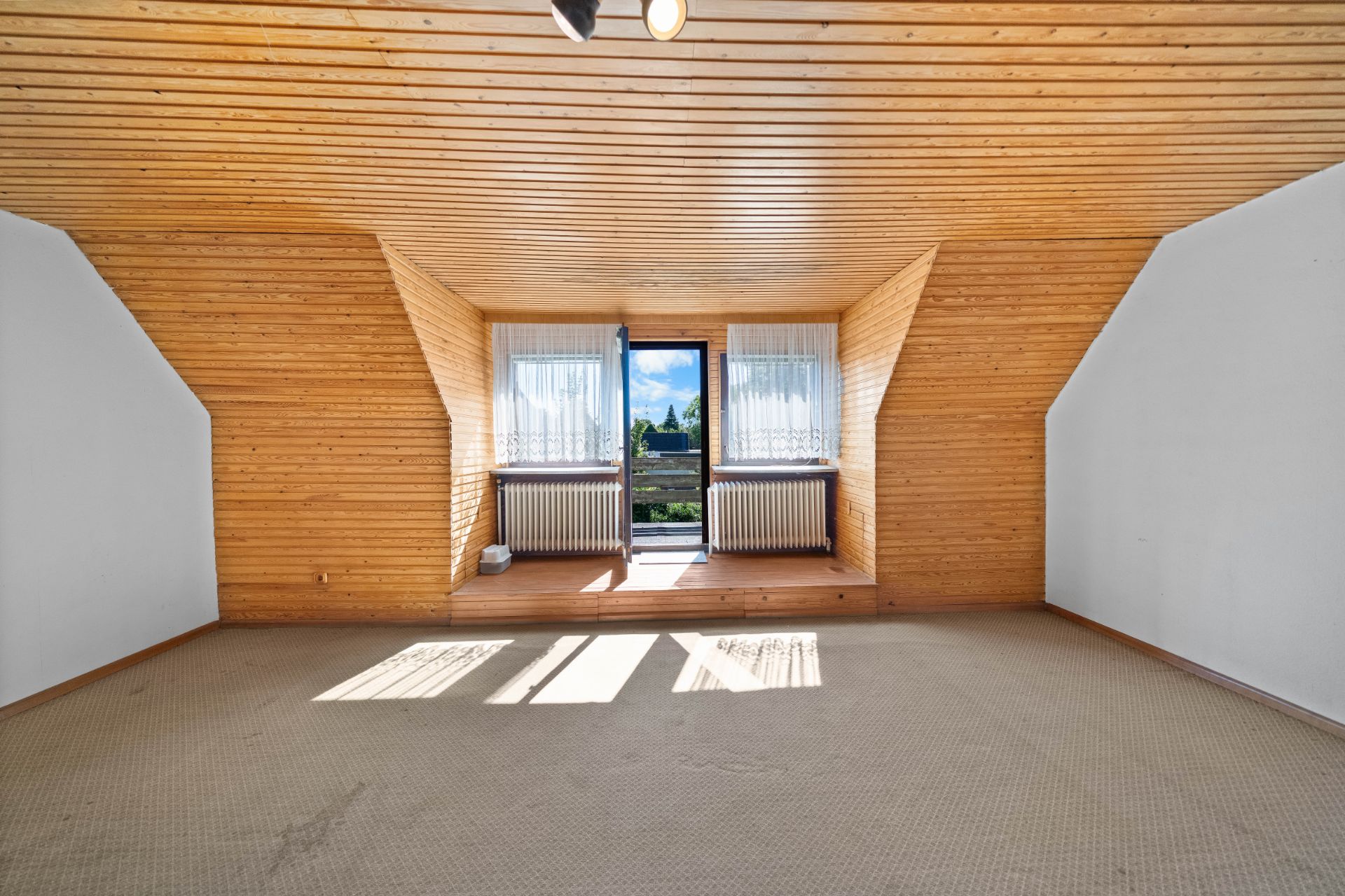 Helles Dachzimmer mit Holzwänden, Teppichboden, zwei Fenstern und einer Glastür zum Balkon.