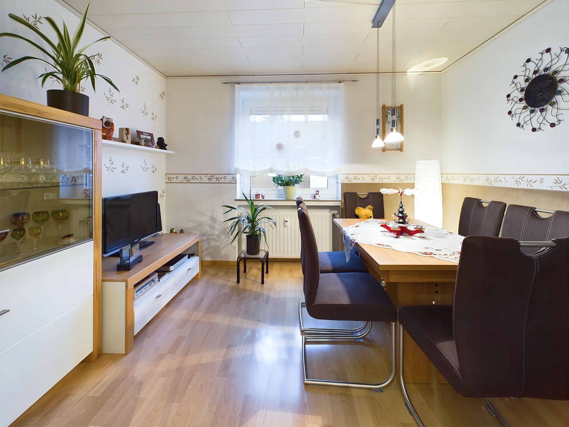 Helles Wohnzimmer mit Esstisch, Stühlen, Fernseher, Pflanzen und Regal.