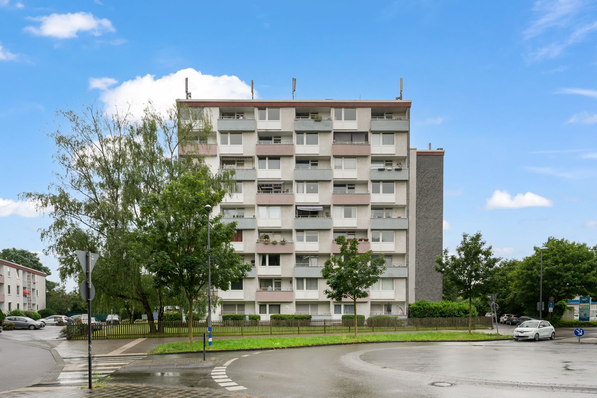 Mehrstöckiges Wohnhaus mit Balkonen und Bäumen davor, an einer nassen Straße gelegen.