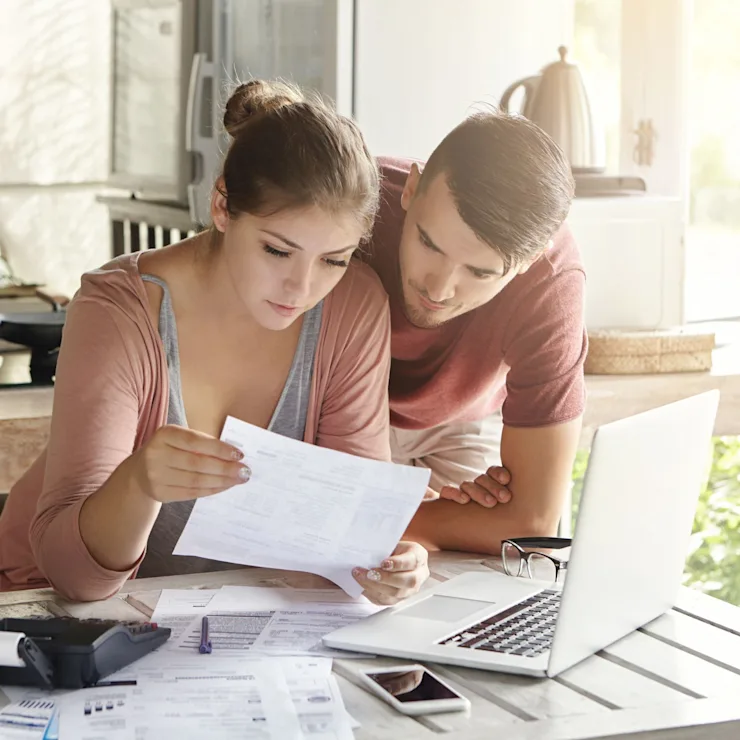 Junges Paar sitzt am Küchentisch und betrachtet gemeinsam Unterlagen mit Laptop und Kaffee.