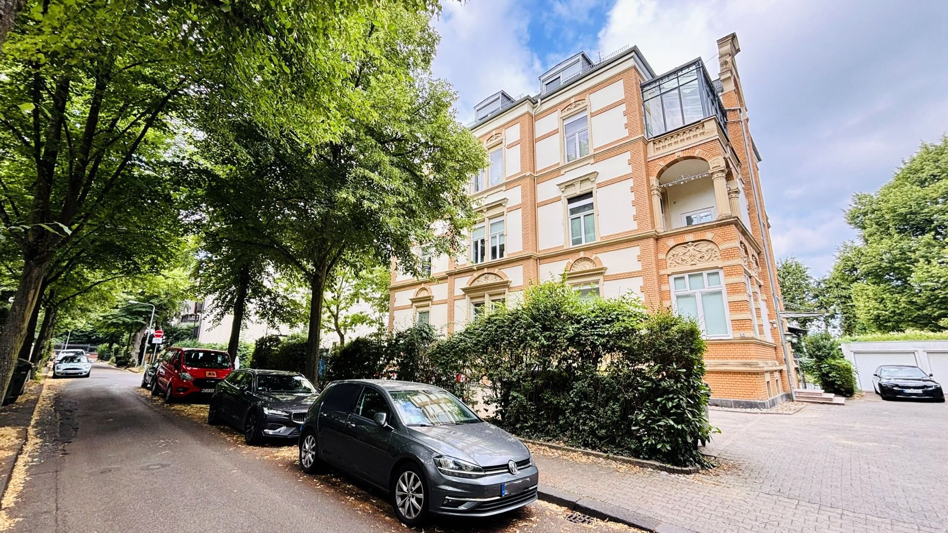 Mehrstöckiges Wohnhaus mit Backsteinfassade an einer baumgesäumten Straße, mehrere Autos parken am Straßenrand.