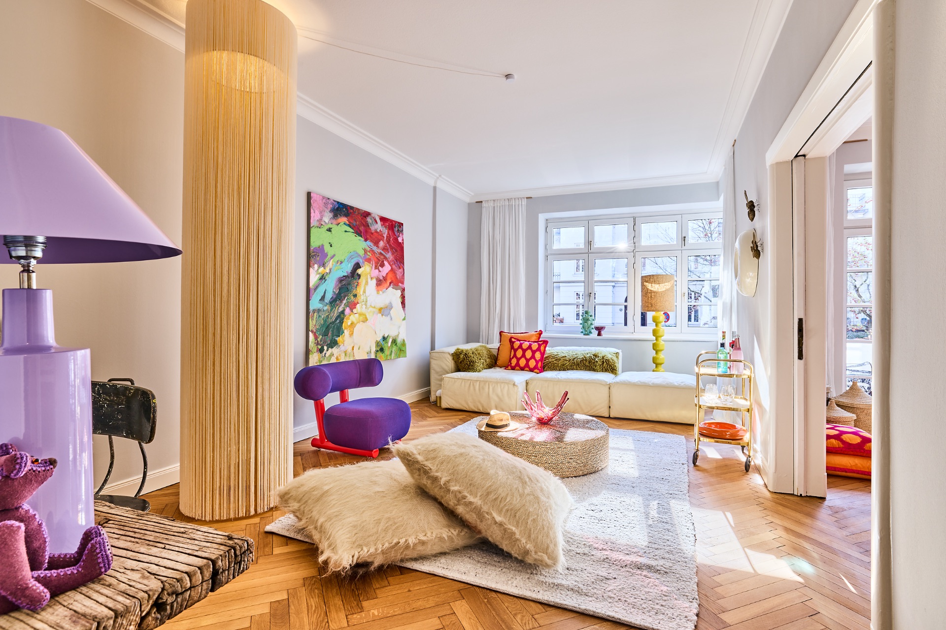Helles Wohnzimmer mit bunten Möbeln, großem Fenster, Teppich und moderner Kunst an der Wand.