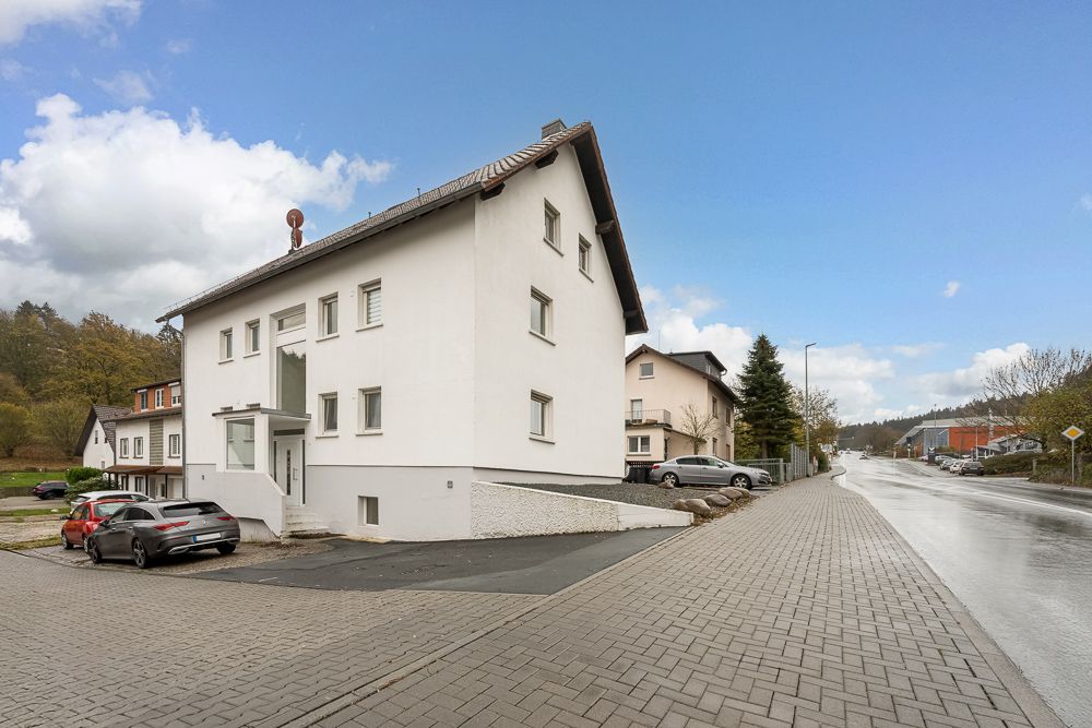 Weißes Wohnhaus mit Satteldach an einer gepflasterten Straße, mehrere Autos parken daneben.