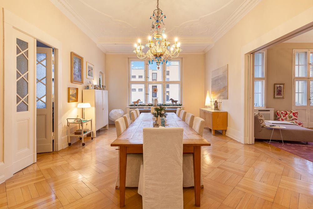 Helles Esszimmer mit langem Holztisch, zwölf Stühlen und Kronleuchter, Parkettboden und großen Fenstern.