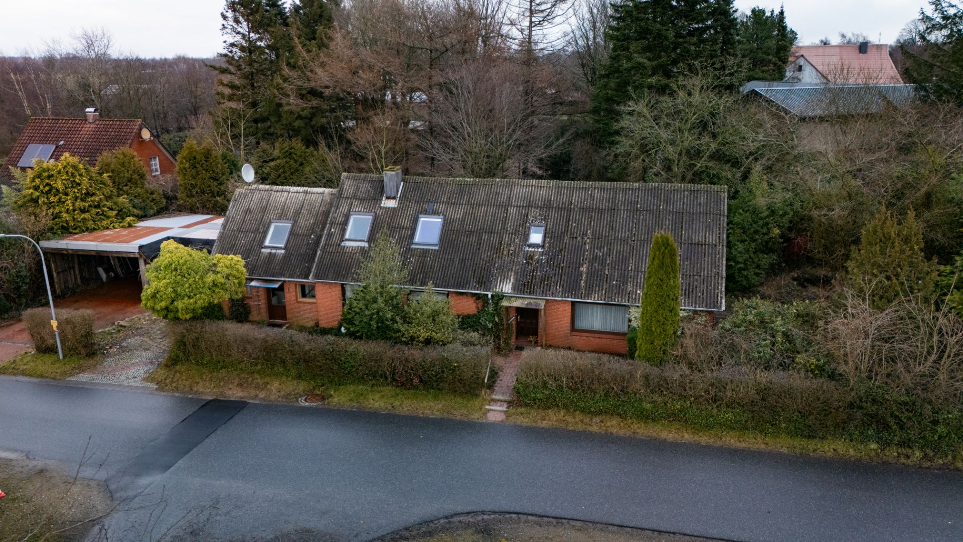 Ziegelhaus mit schrägem Dach, mehreren Dachfenstern und Büschen vor dem Haus an einer Straße.