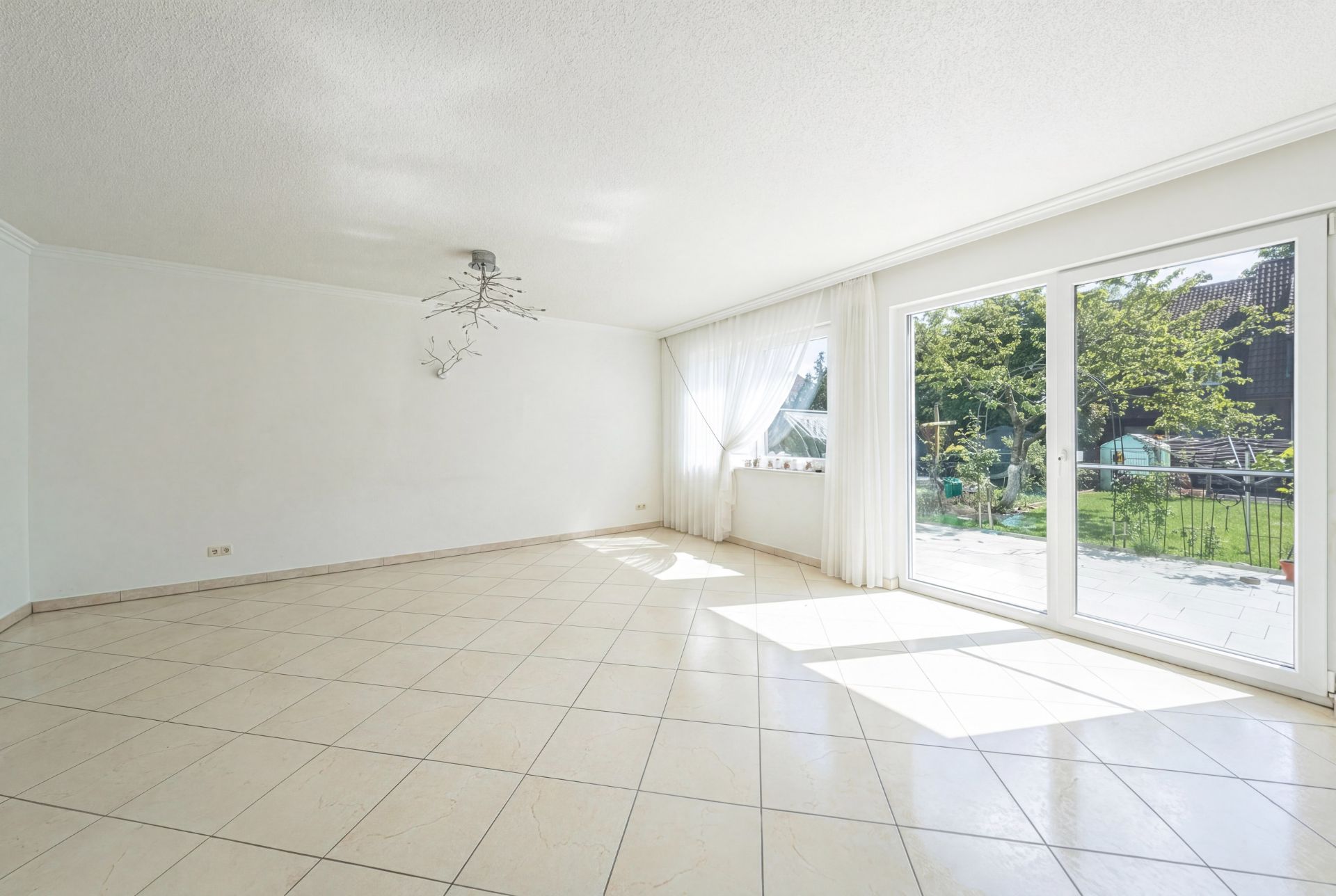 Heller leerer Raum mit Fliesenboden, großem Fenster und Blick in den Garten.