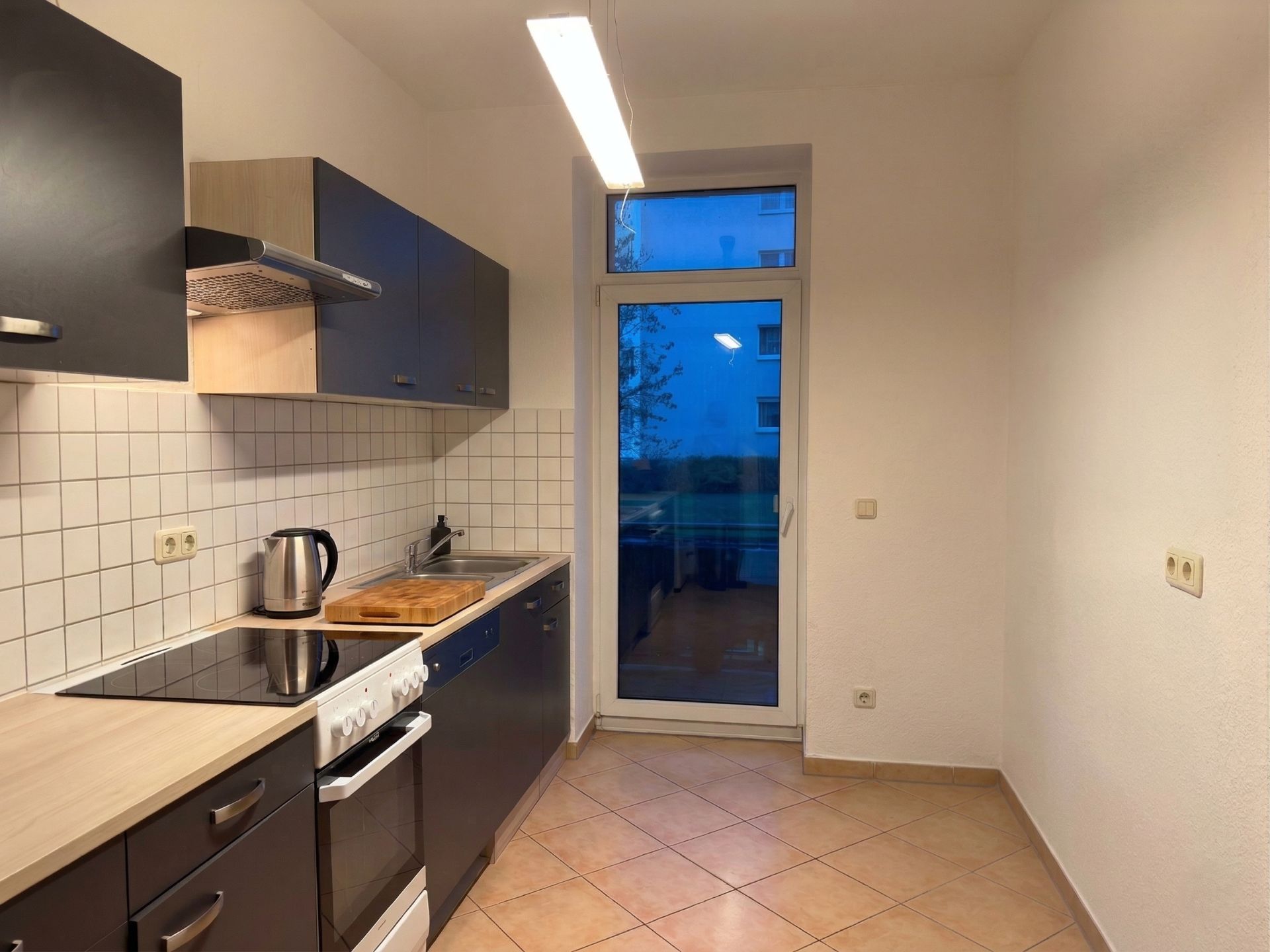 Moderne Küche mit Fliesenboden, Herd, Spüle, Hängeschränken und Glastür zum Garten.