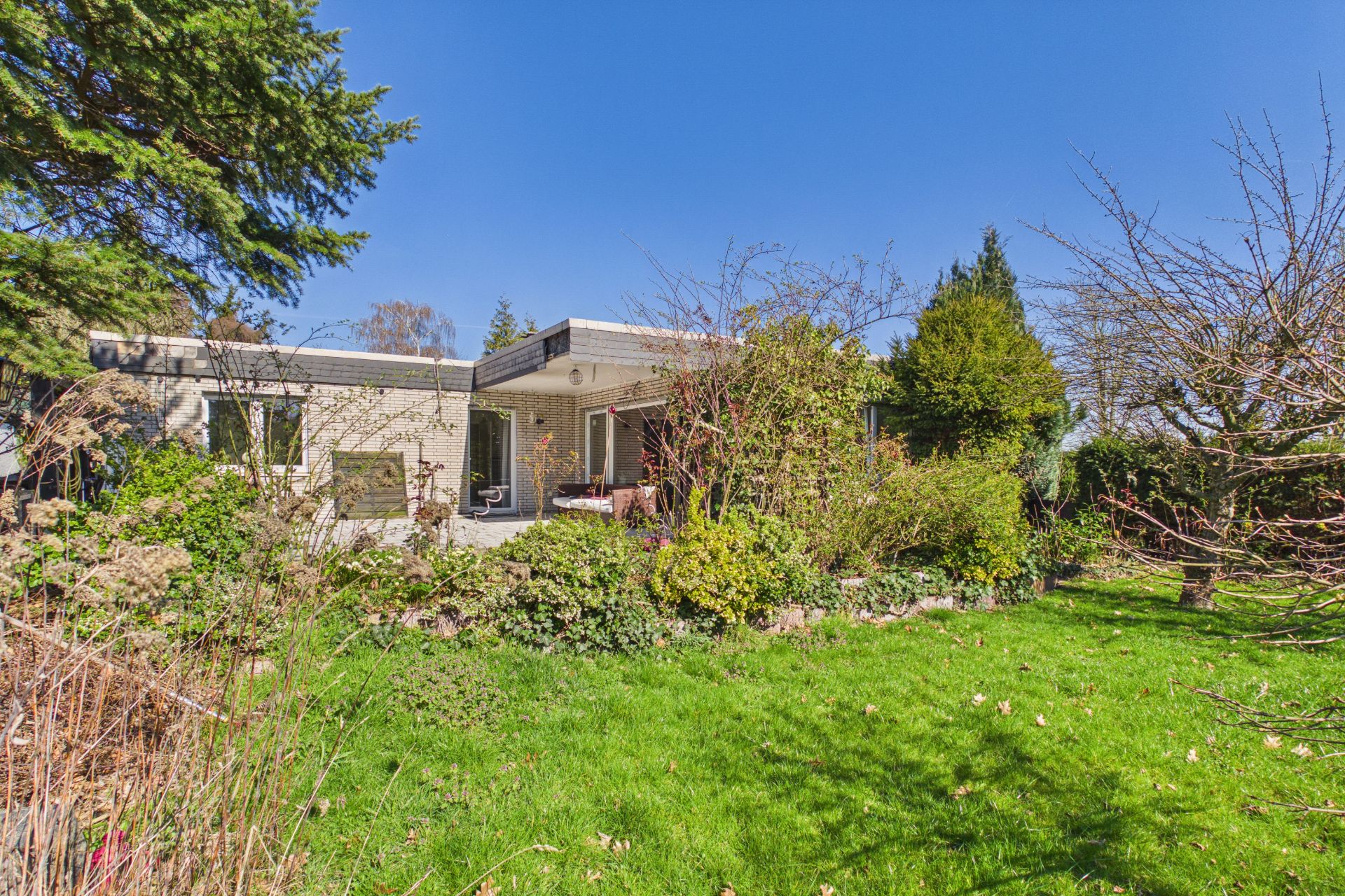 Flaches Einfamilienhaus mit Terrasse und großem Garten, viele Sträucher und Bäume, blauer Himmel.