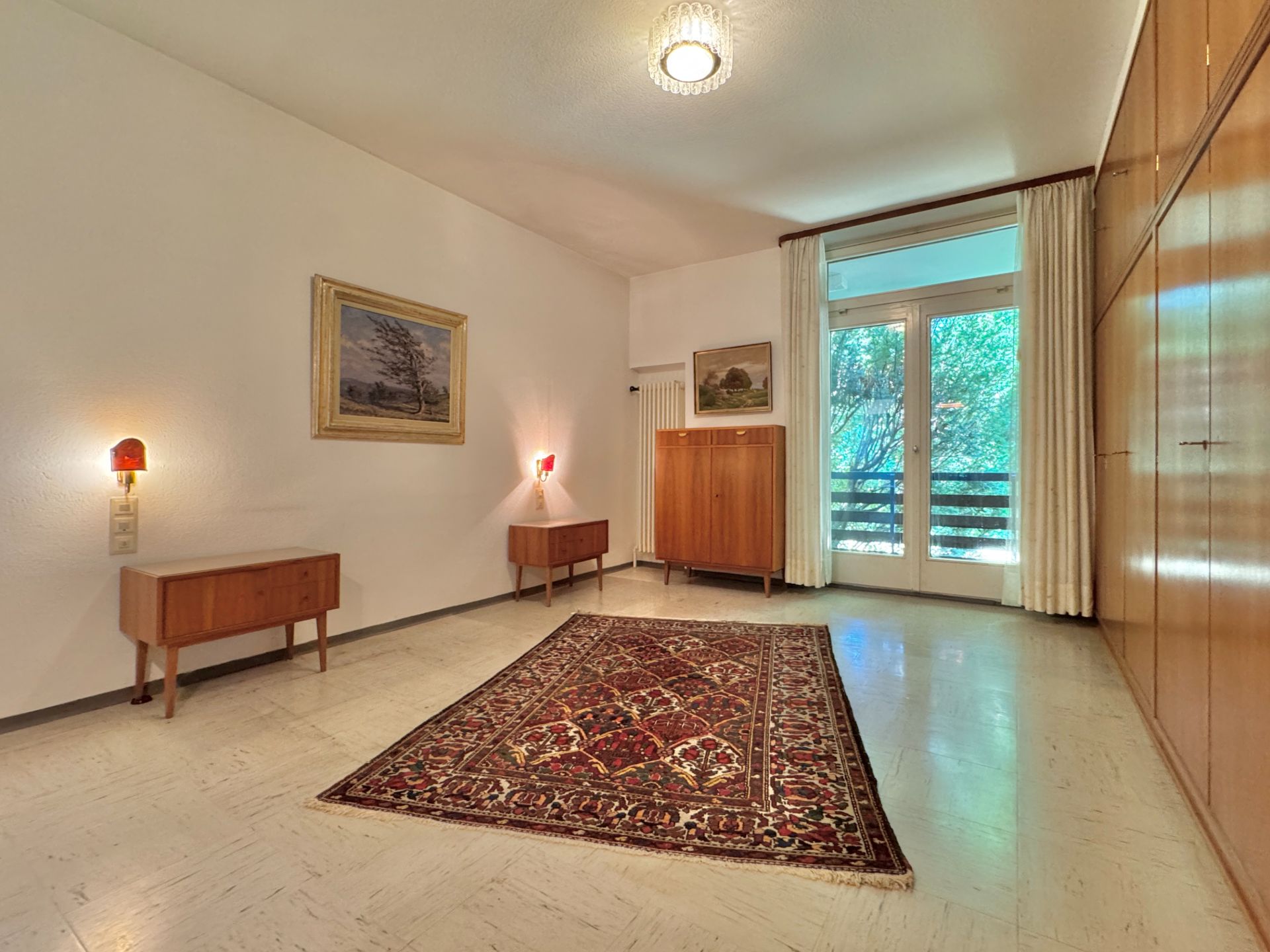 Helles Wohnzimmer mit Teppich, Holzmöbeln, Wandbildern und großem Fenster mit Vorhängen.