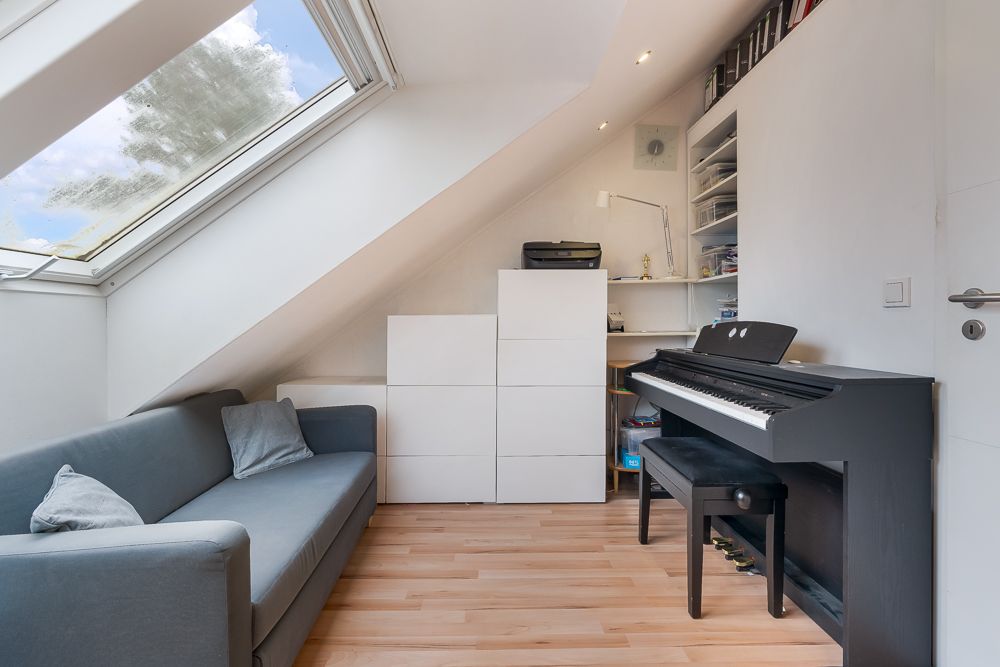 Dachzimmer mit Sofa, Klavier, Regalen und großem Dachfenster.