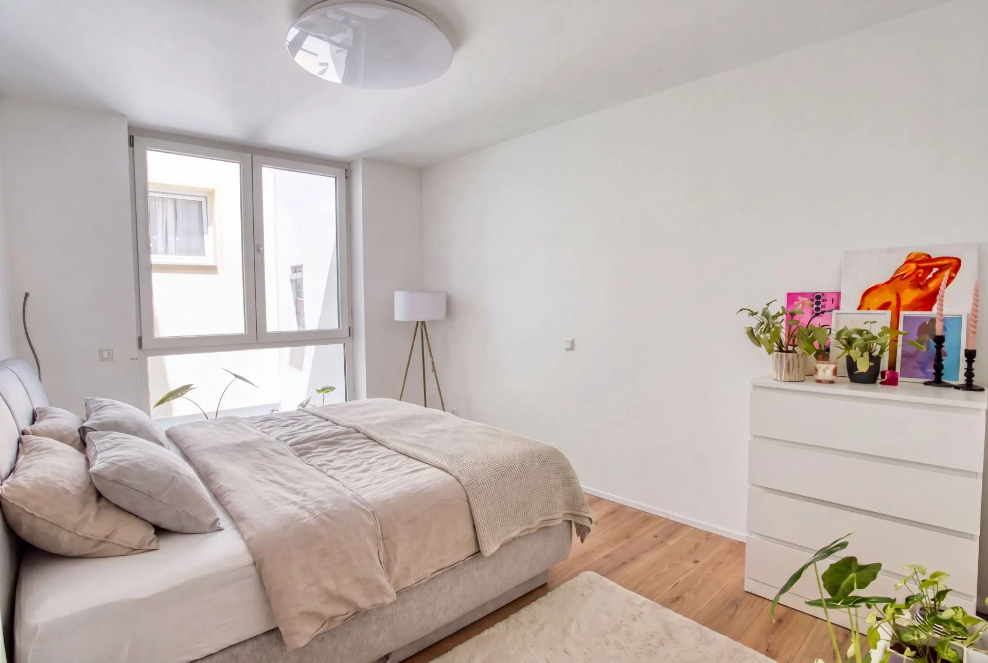 Helles Schlafzimmer mit Doppelbett, Stehlampe, Kommode und Pflanzen auf Holzfußboden.
