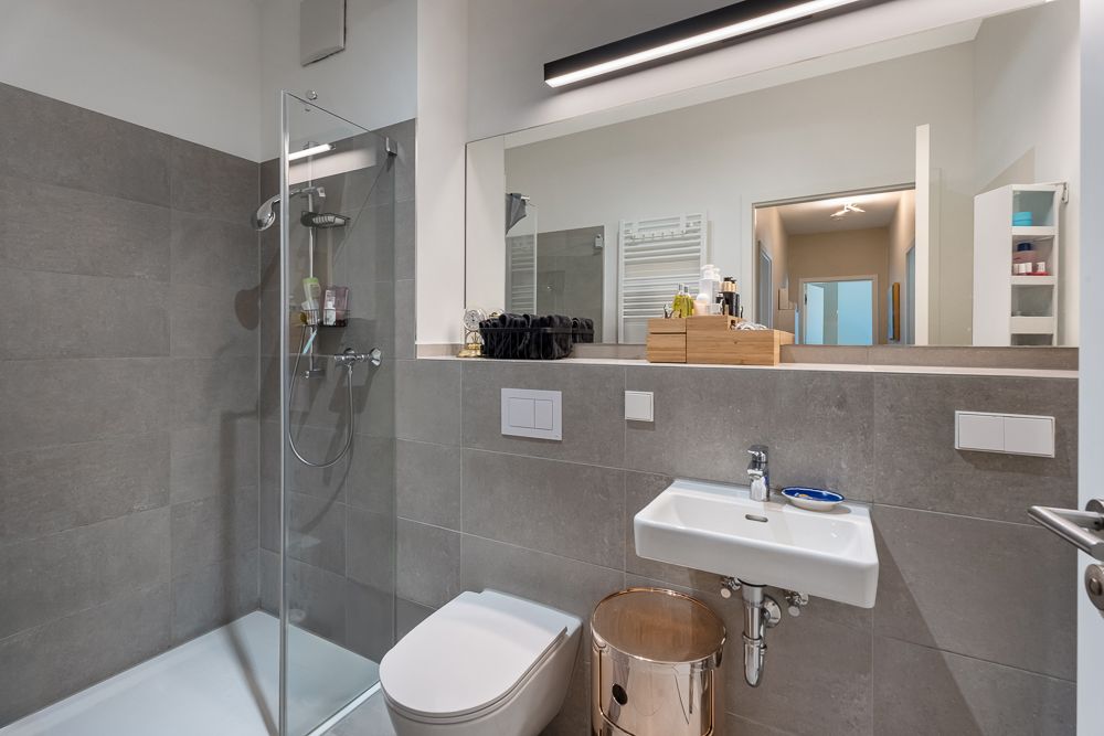 Modernes Badezimmer mit grauen Fliesen, Dusche, Waschbecken, WC und großem Spiegel.