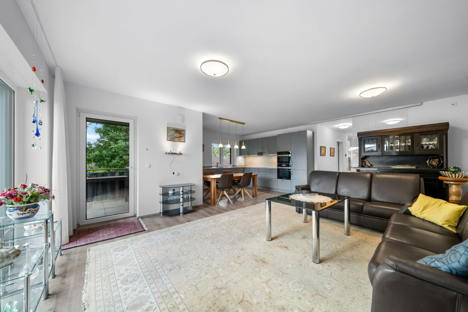Wohnzimmer mit Ledersofa, Glastisch, Teppich, Essbereich und moderner Küche, Zugang zum Balkon.
