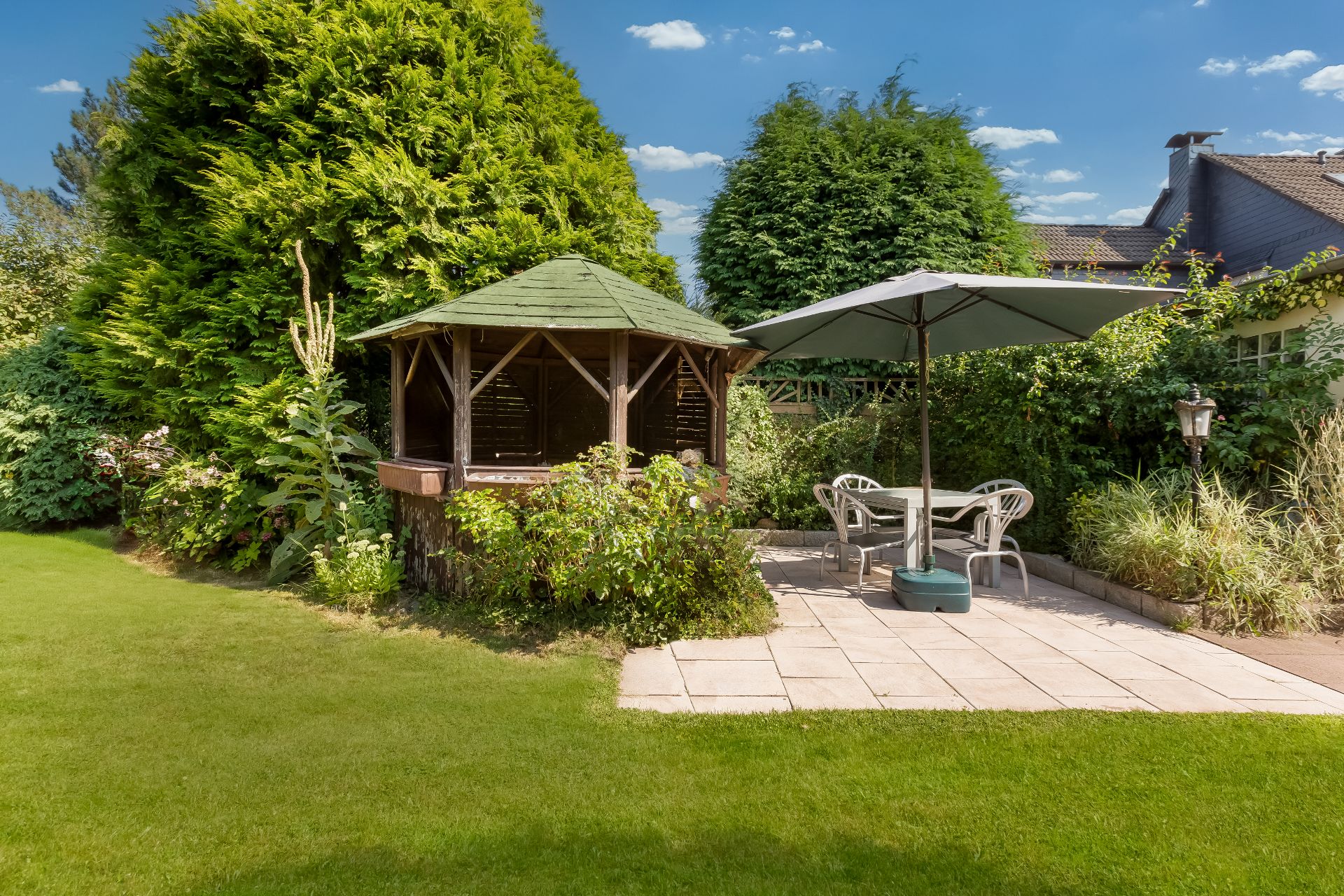 Garten mit Holzpavillon, Terrasse, Tisch, Stühlen und großem Sonnenschirm.