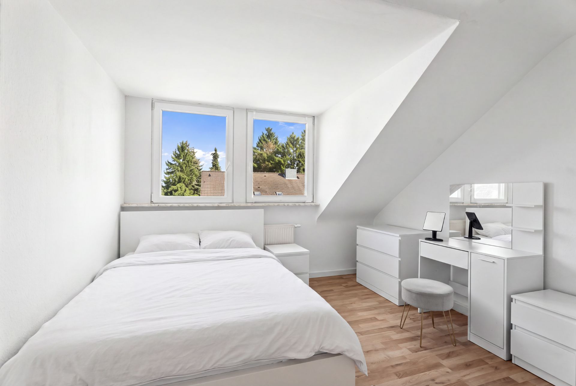 Helles Schlafzimmer mit Bett, Kommode, Schminktisch und zwei Fenstern, Blick ins Grüne.