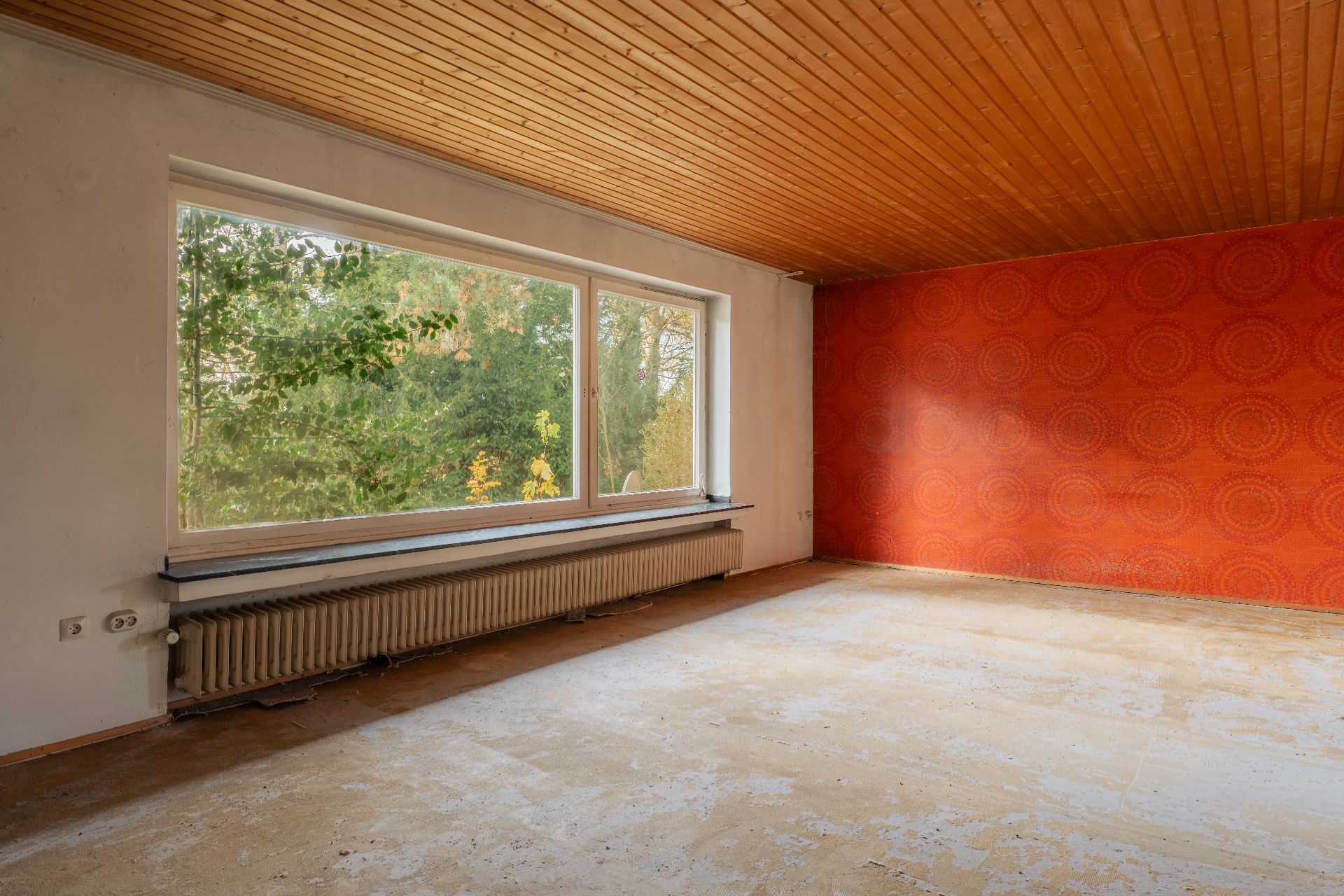 Leerer Raum mit großem Fenster, Heizung, Holzdecke und roter Musterwand.