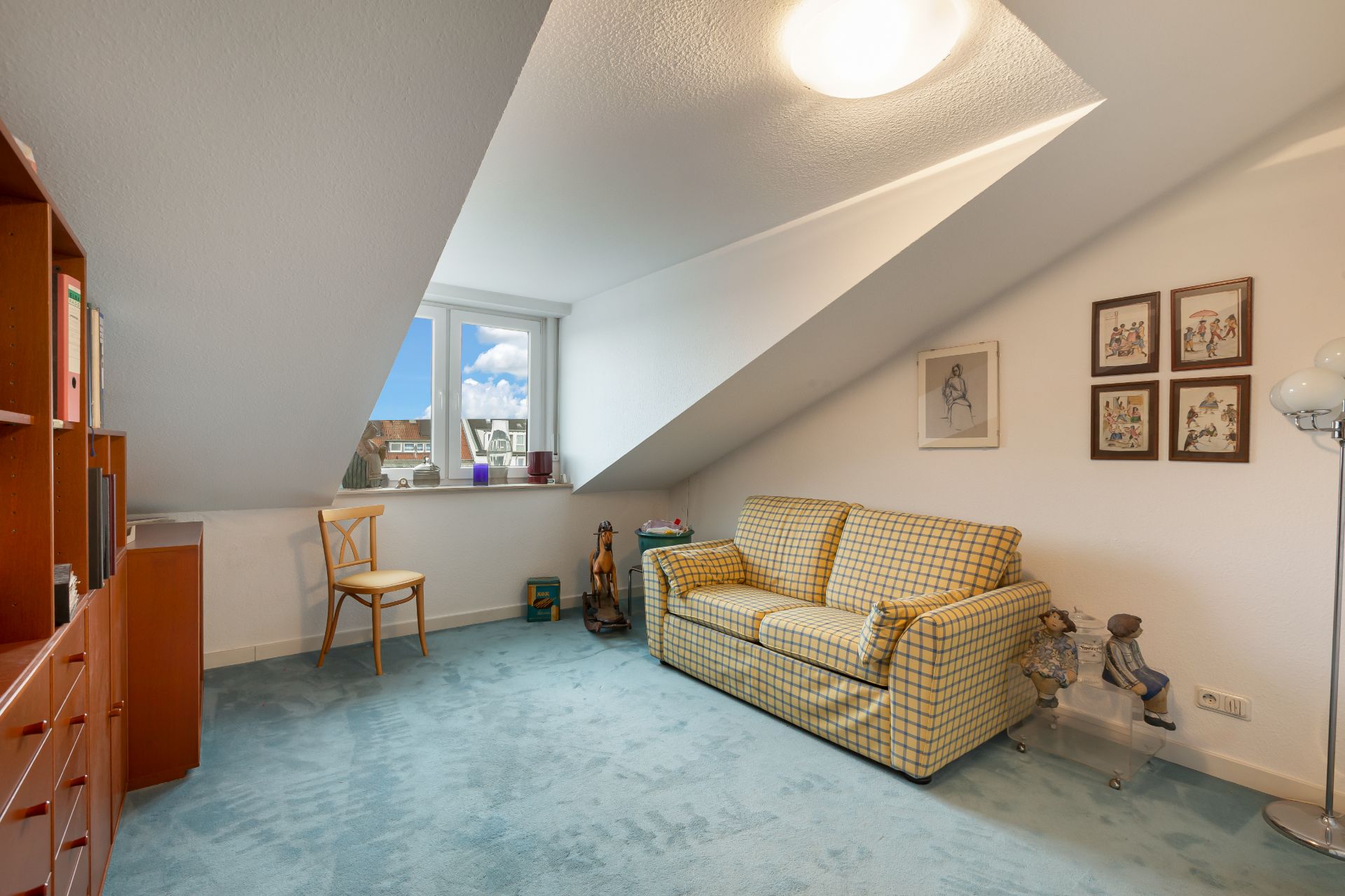 Dachzimmer mit Sofa, blauem Teppich, Regal, Stuhl und schräger Decke.