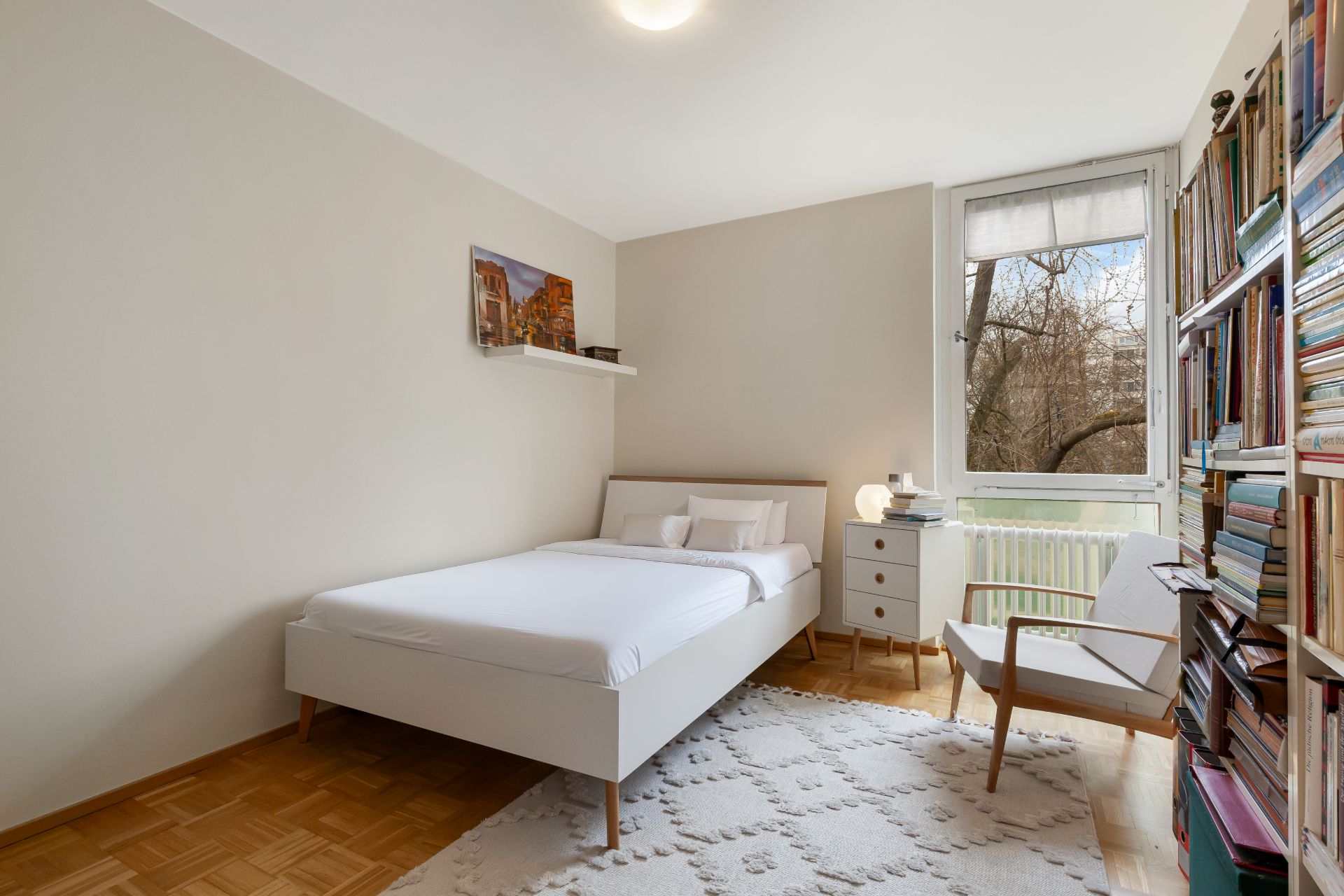 Helles Schlafzimmer mit Doppelbett, Bücherregal, Sessel und großem Fenster.