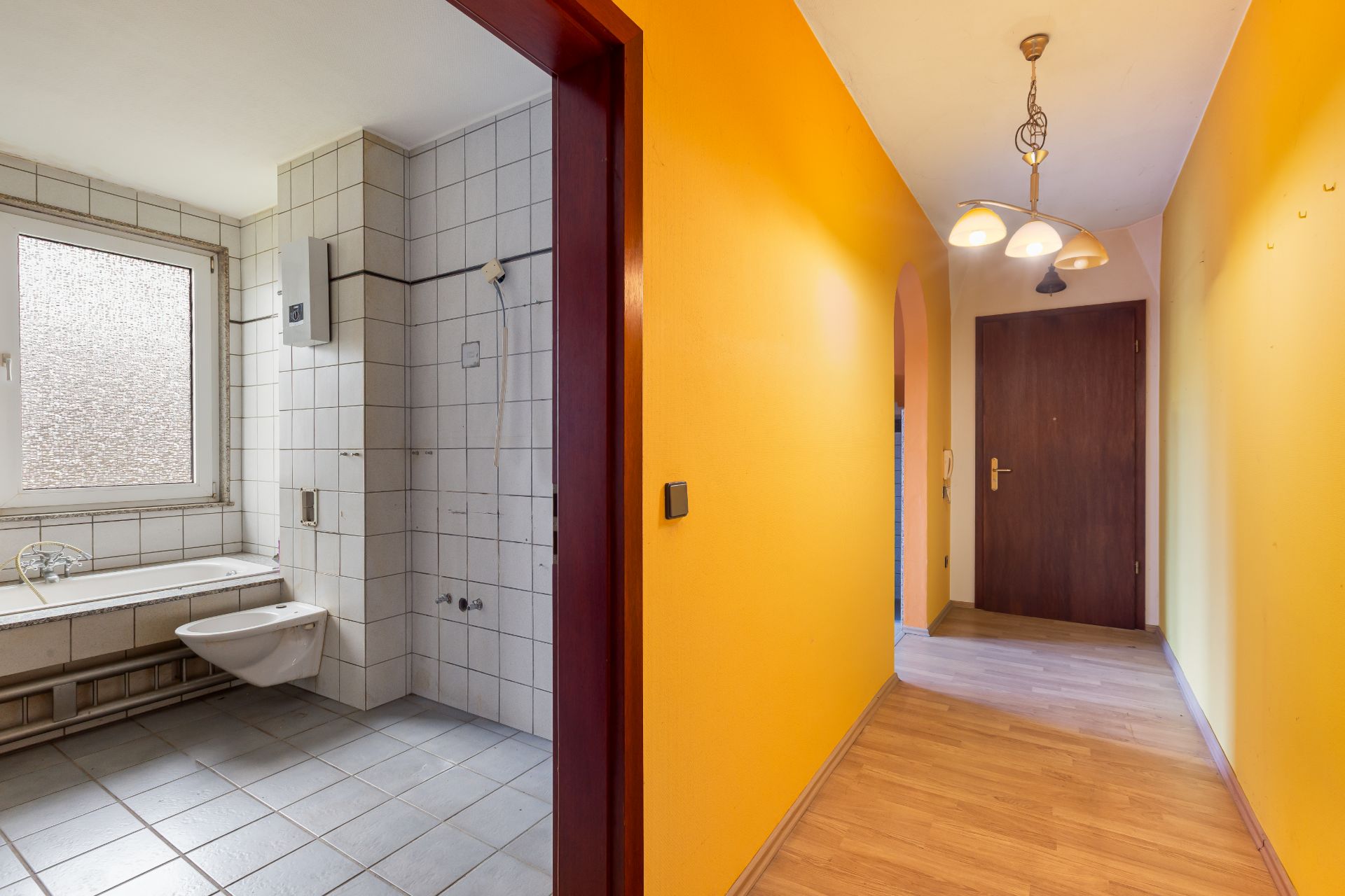Flur mit gelben Wänden, rechts eine Tür, links ein Badezimmer mit weißen Fliesen und Fenster.