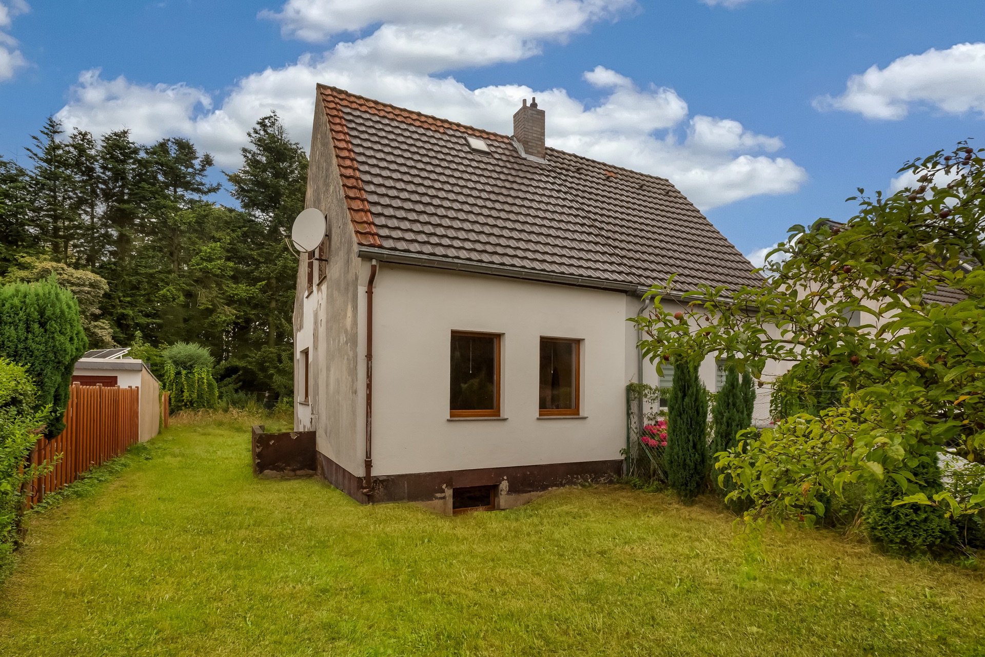 Ein kleines Einfamilienhaus mit Garten, Bäumen und einem Holzzaun an der Seite.