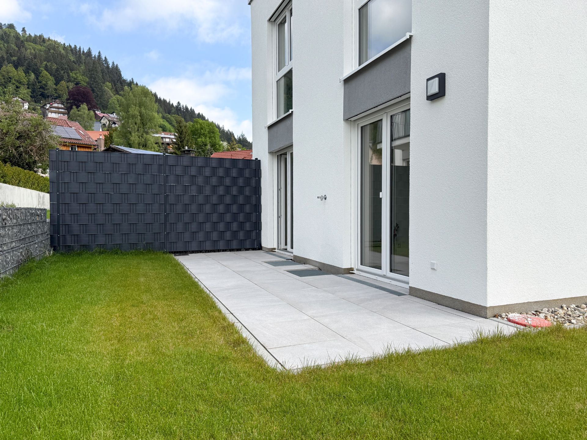 Moderne Terrasse mit Fliesenboden und Glastüren an einem weißen Wohnhaus mit kleinem Garten.