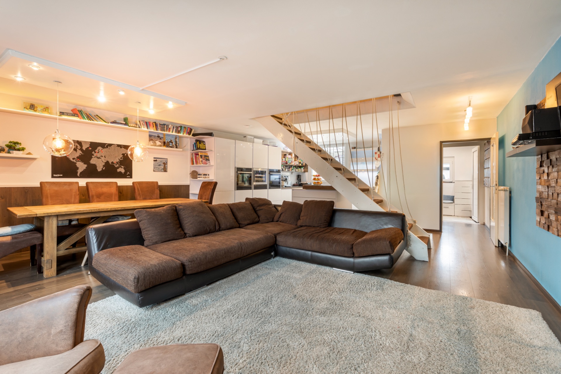 Modernes Wohnzimmer mit braunem Ecksofa, Esstisch, offener Küche und Treppe nach oben.