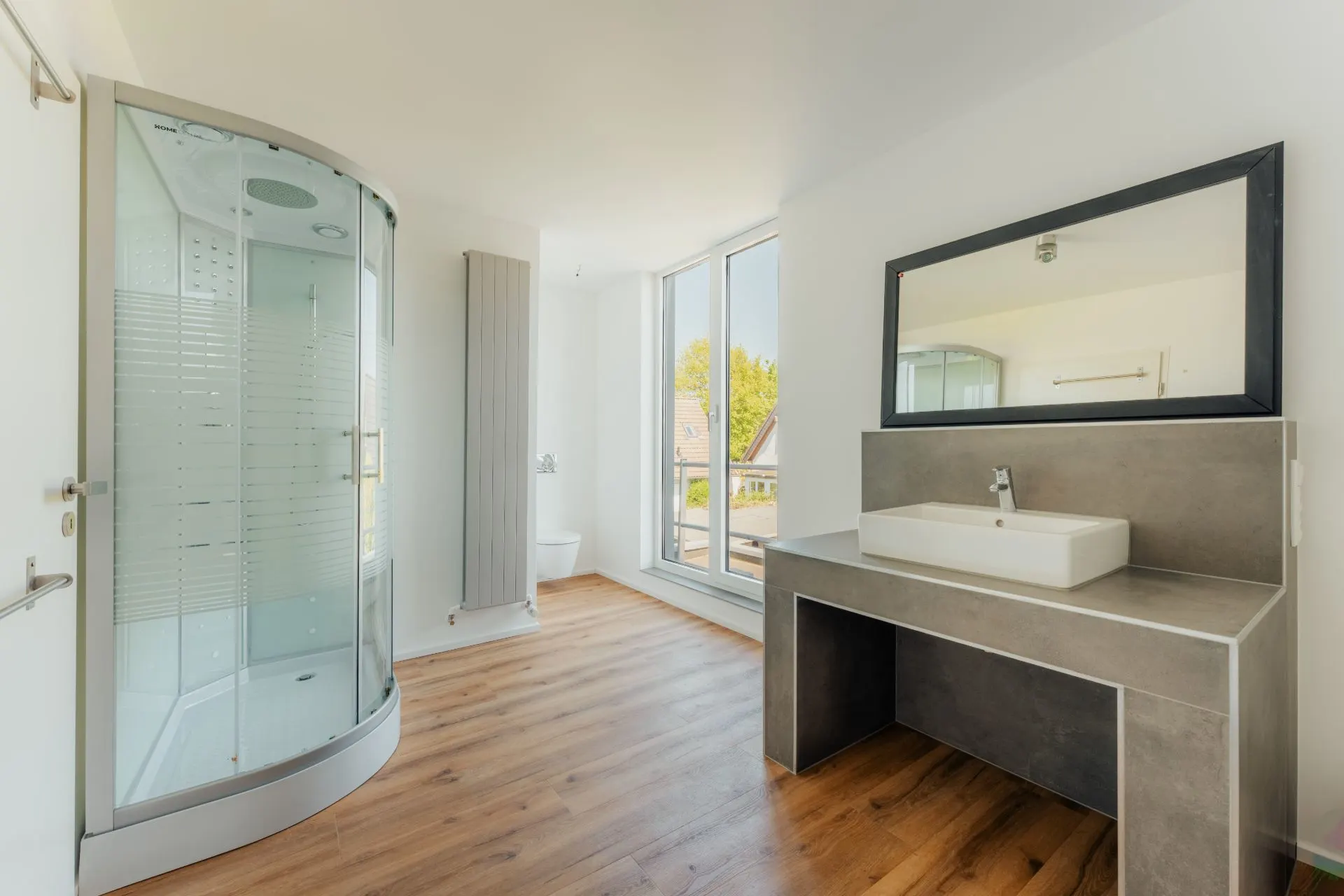Helles Badezimmer mit Dusche, Waschbecken, großem Spiegel und Fenster, Parkettboden.