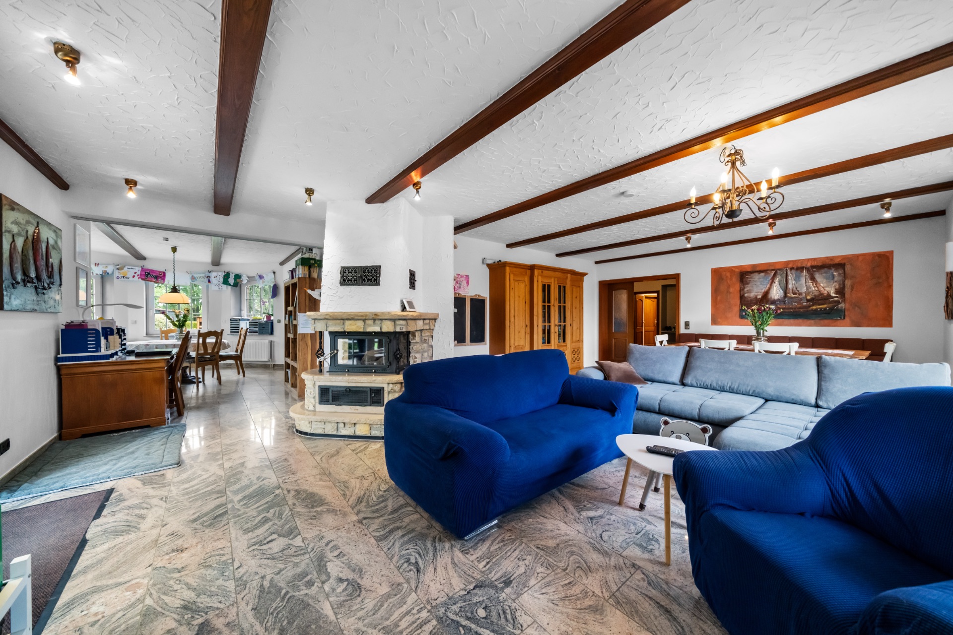 Wohnzimmer mit blauen Sofas, Kamin, Esstisch und Fenster, offene Verbindung zur Küche.