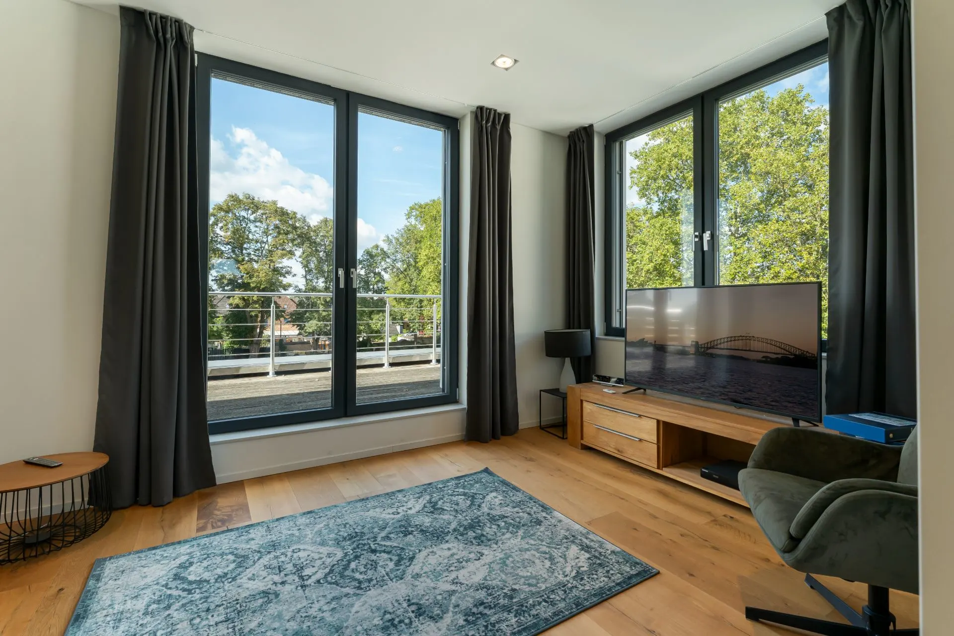 Wohnzimmer mit großem Fenster, grauen Vorhängen, Teppich, Fernseher und Holzfußboden.
