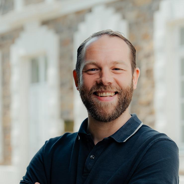 Ein Mann mit Bart steht draußen vor einem Gebäude und lächelt mit verschränkten Armen.