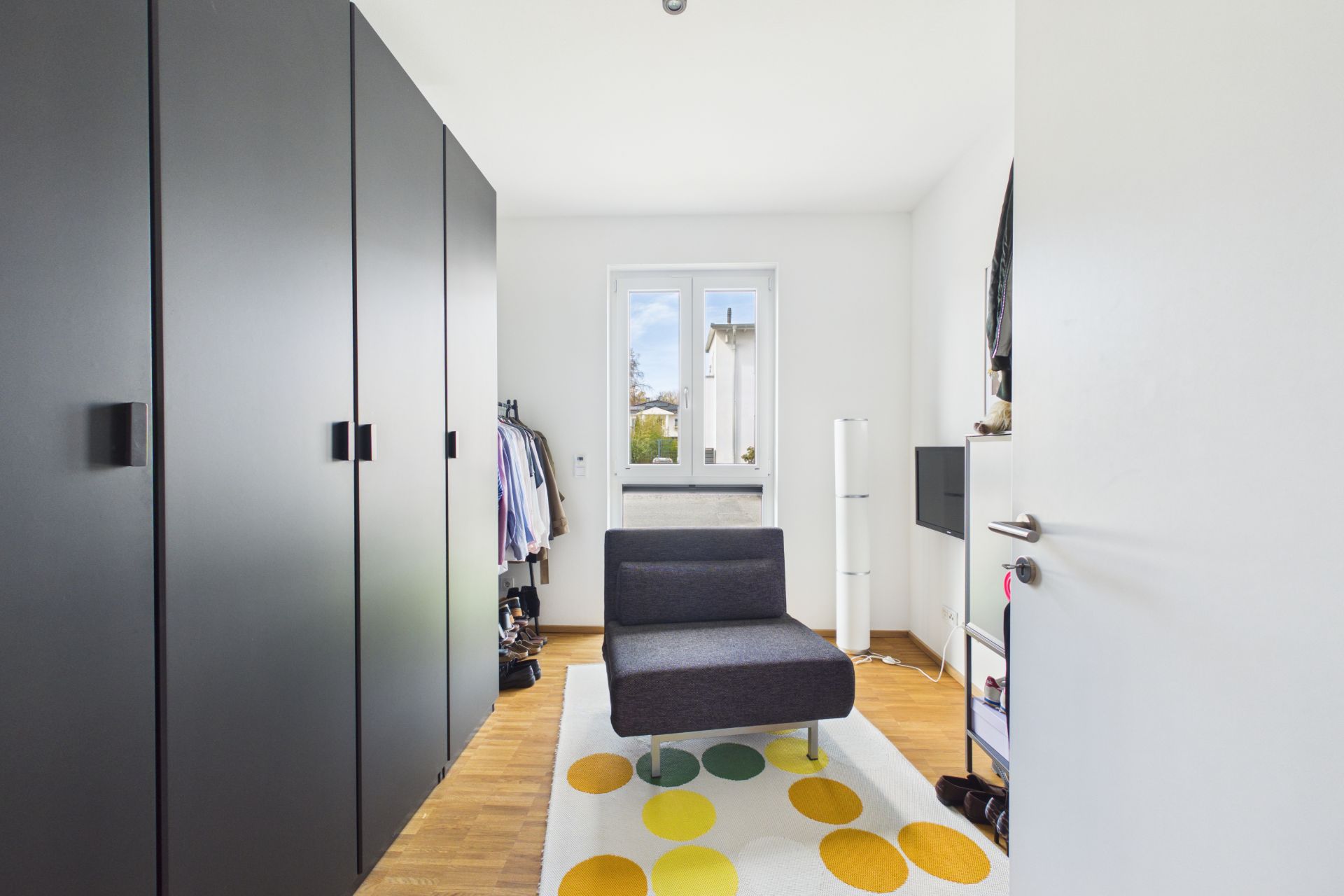 Kleines Zimmer mit schwarzem Sessel, Kleiderschränken und Teppich mit bunten Kreisen.