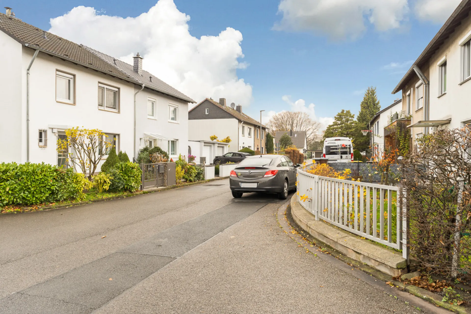 Wohnstraße mit geparktem Auto, weißen Häusern und Vorgärten an einem bewölkten Tag.