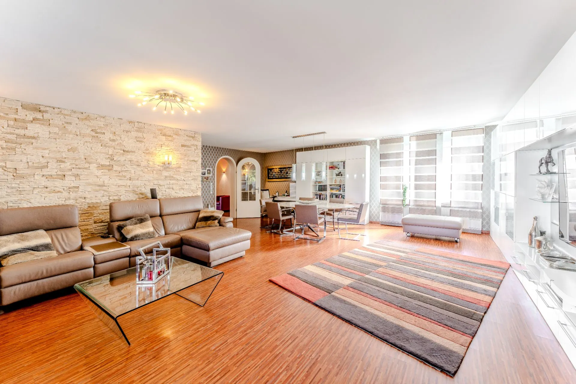 Helles Wohnzimmer mit Ledersofas, Glastisch, Teppich, Essbereich und großen Fenstern.
