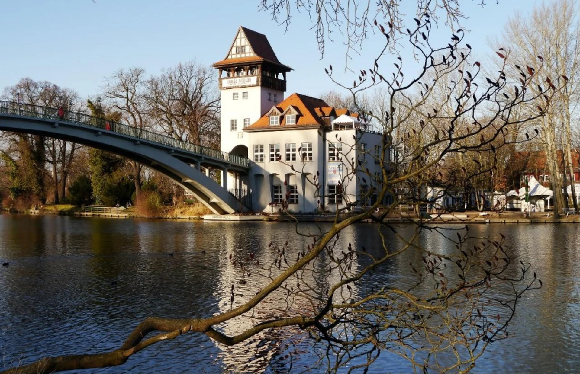 Großes Haus mit Turm am Fluss, daneben eine Brücke und Bäume im Vordergrund.