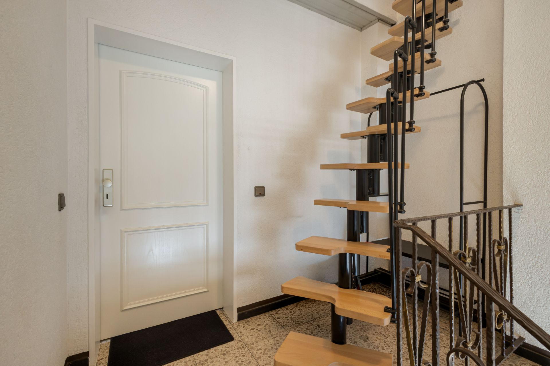 Innenraum mit weißer Tür und einer offenen Holztreppe mit Metallgeländer.