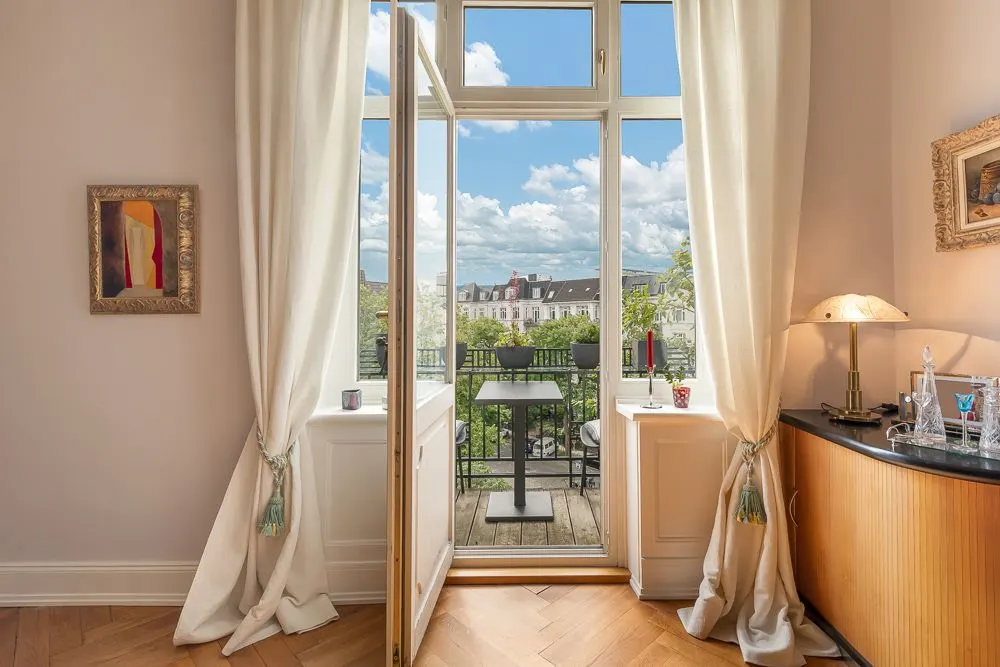 Offene Balkontür mit Ausblick auf einen Balkon, einen Tisch und Wohnhäuser im Hintergrund.